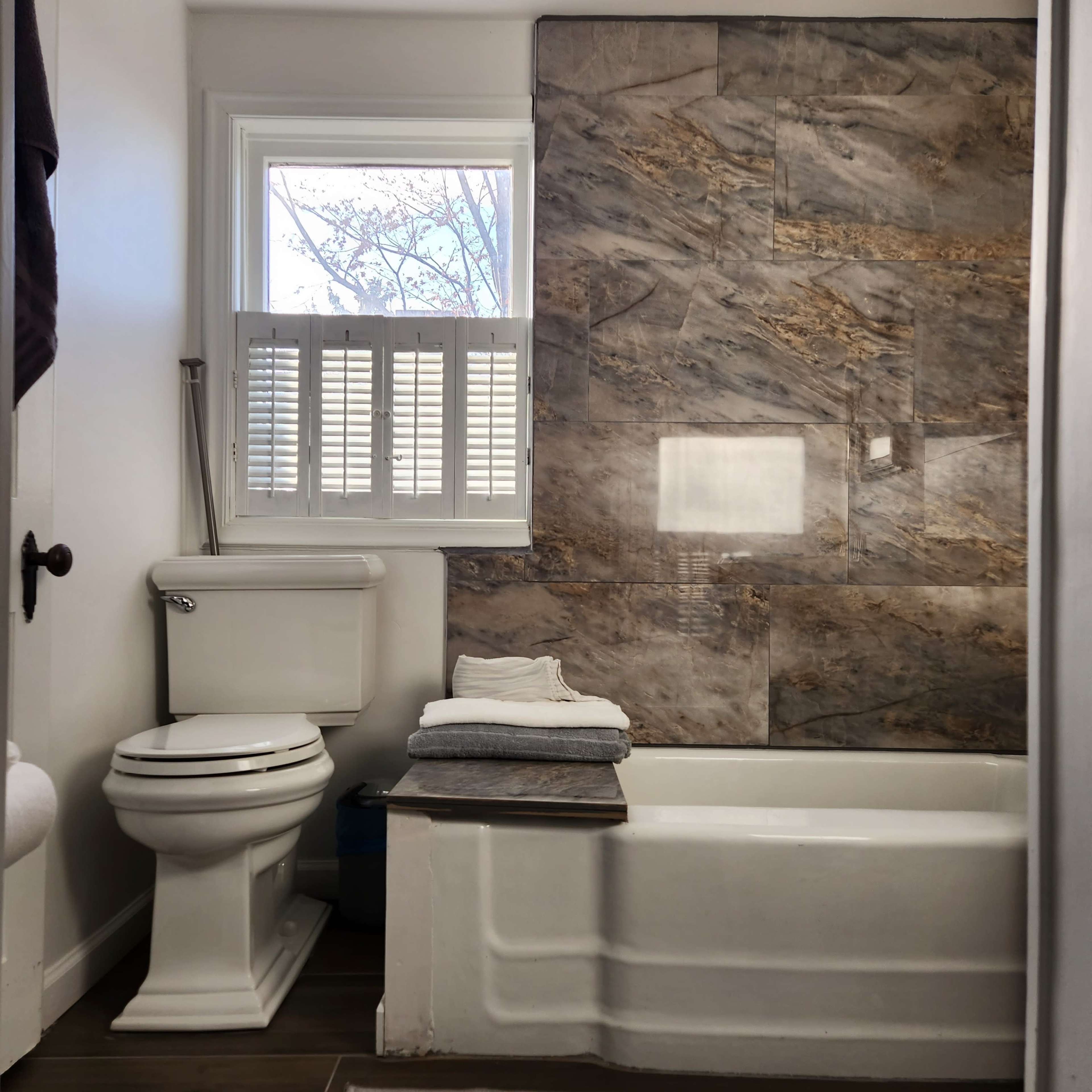 A clean bathroom featuring a white toilet, a bathtub with a wooden seat, and a marble wall with a window above.