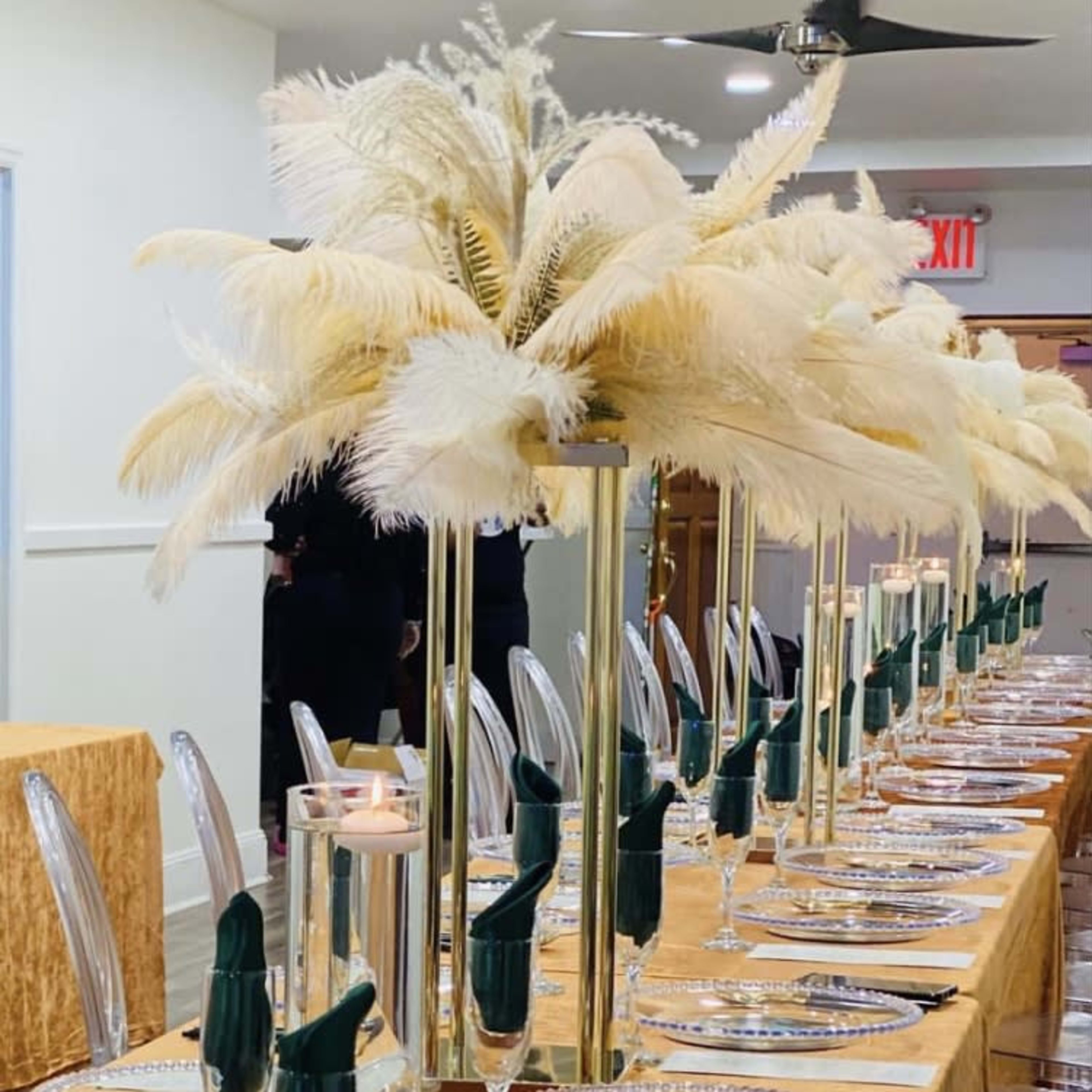 A long dining table is elegantly set with gold tablecloths, transparent plates, and tall centerpieces featuring white feathers.