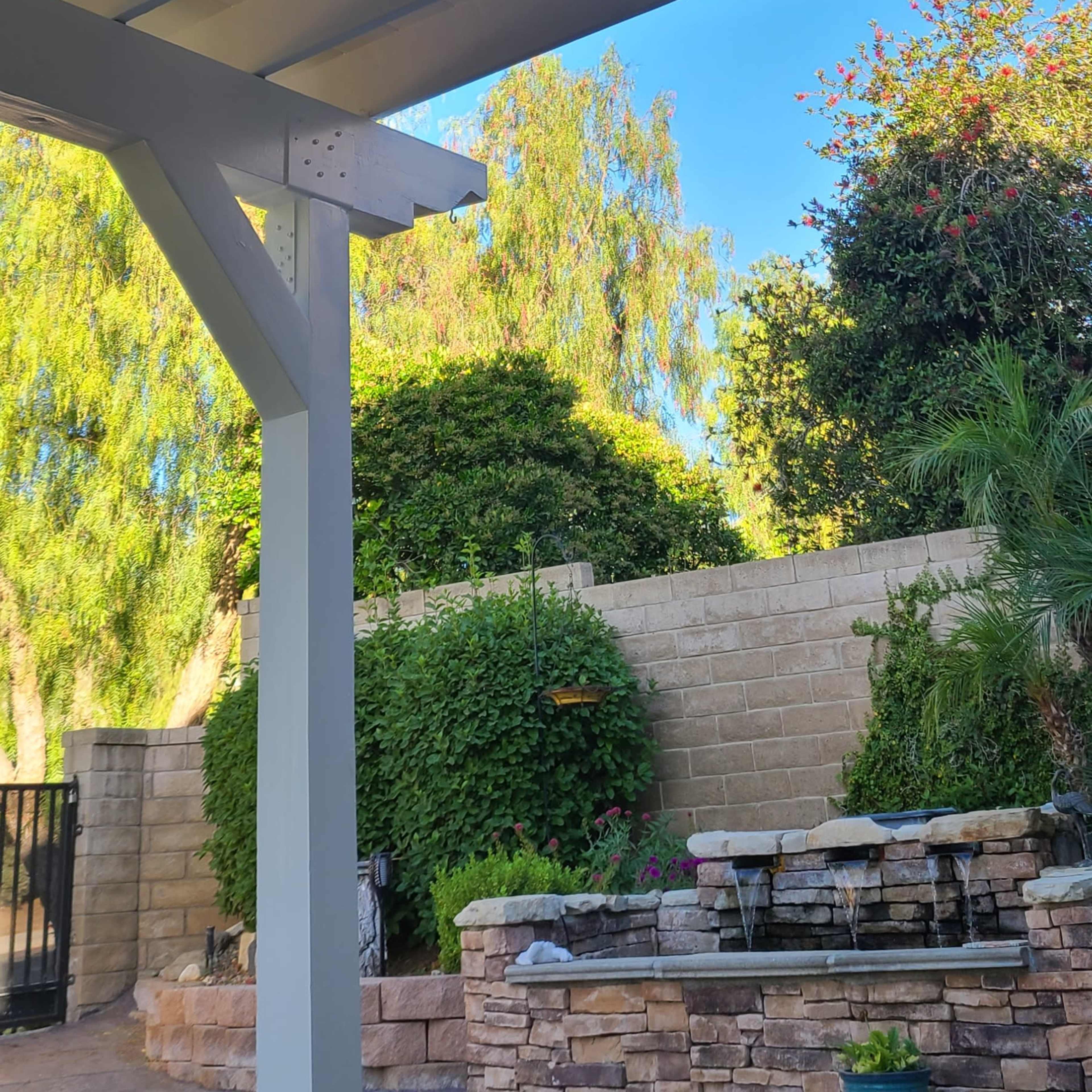 A ceiling fan with five wooden blades and a light fixture is mounted under a covered patio surrounded by greenery.