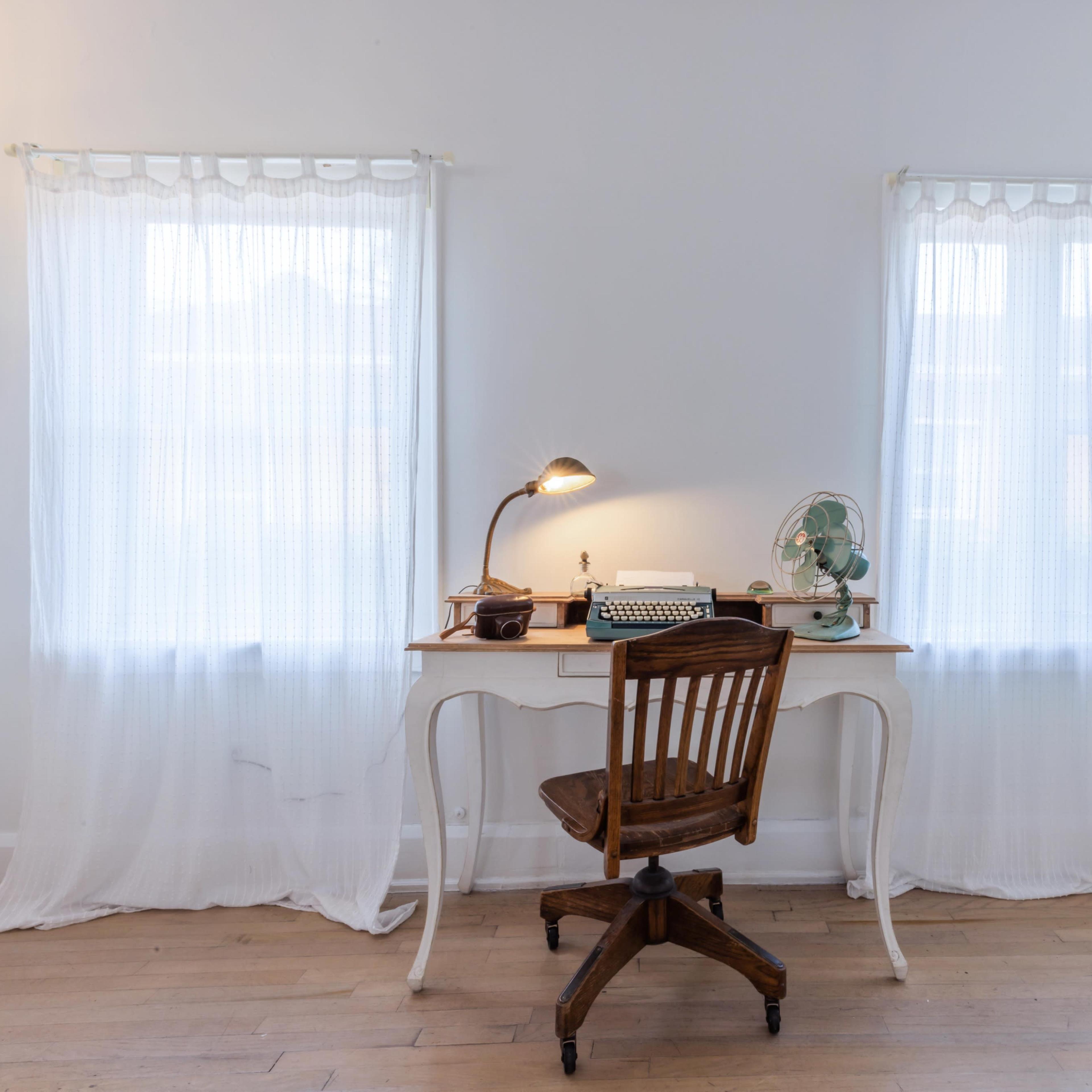 A wooden desk with a chair, a typewriter, and a lamp is positioned between two sheer-curtained windows in a well-lit room.