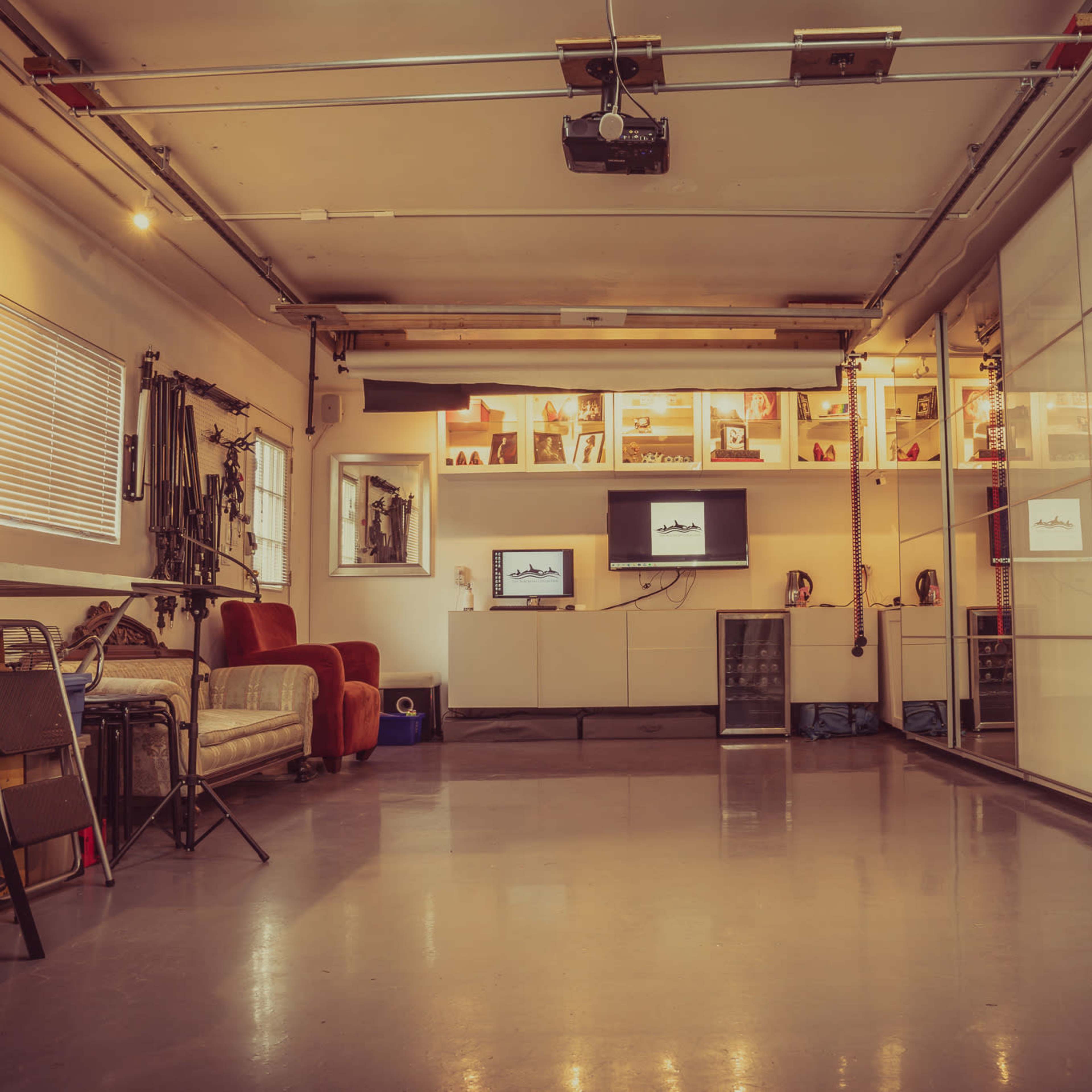 A spacious indoor garage area with a polished floor, fitted with shelves displaying various items, a projector on the ceiling, and two televisions mounted on a wall.