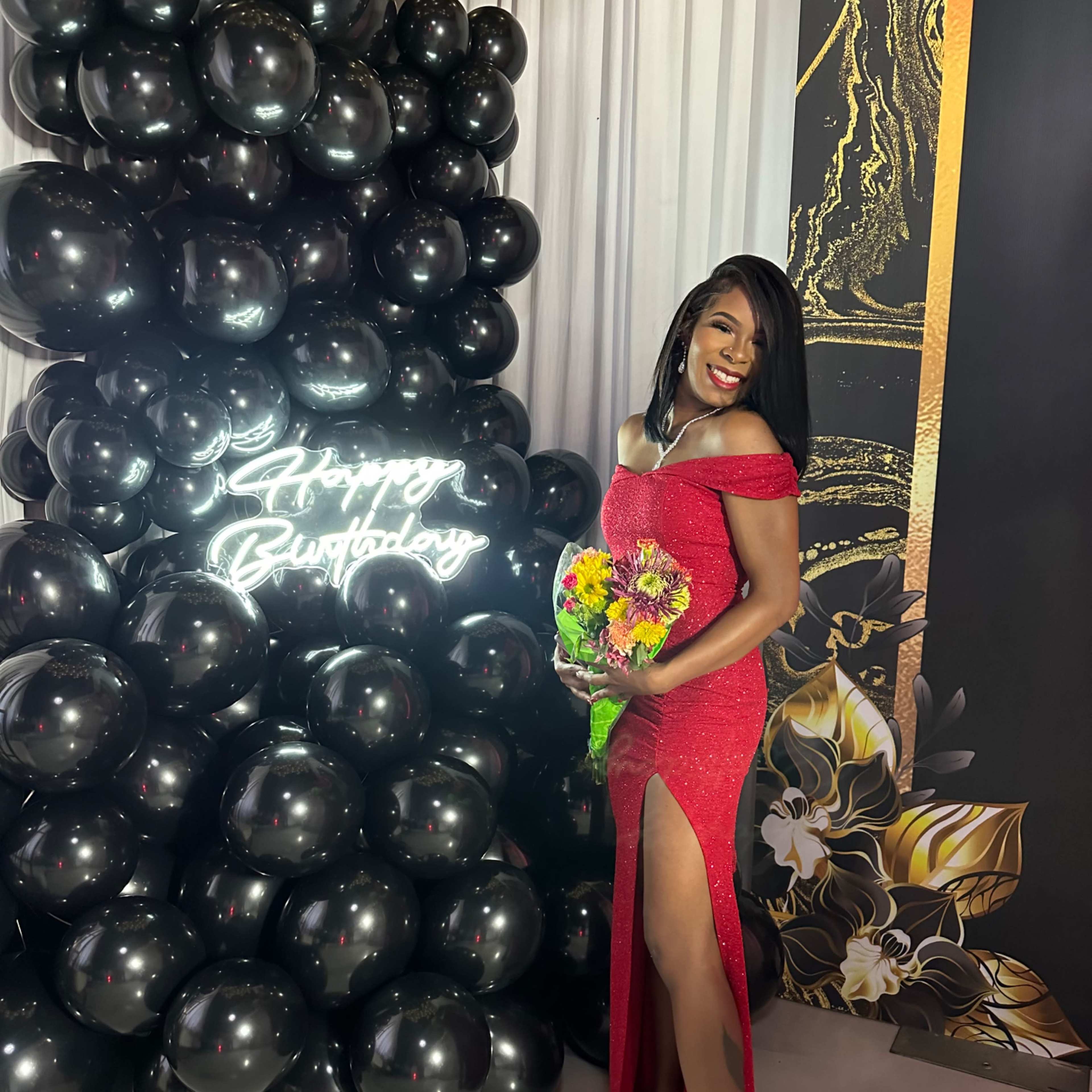 A woman in a red dress with a thigh-high slit poses next to a black balloon backdrop featuring a "Happy Birthday" sign.