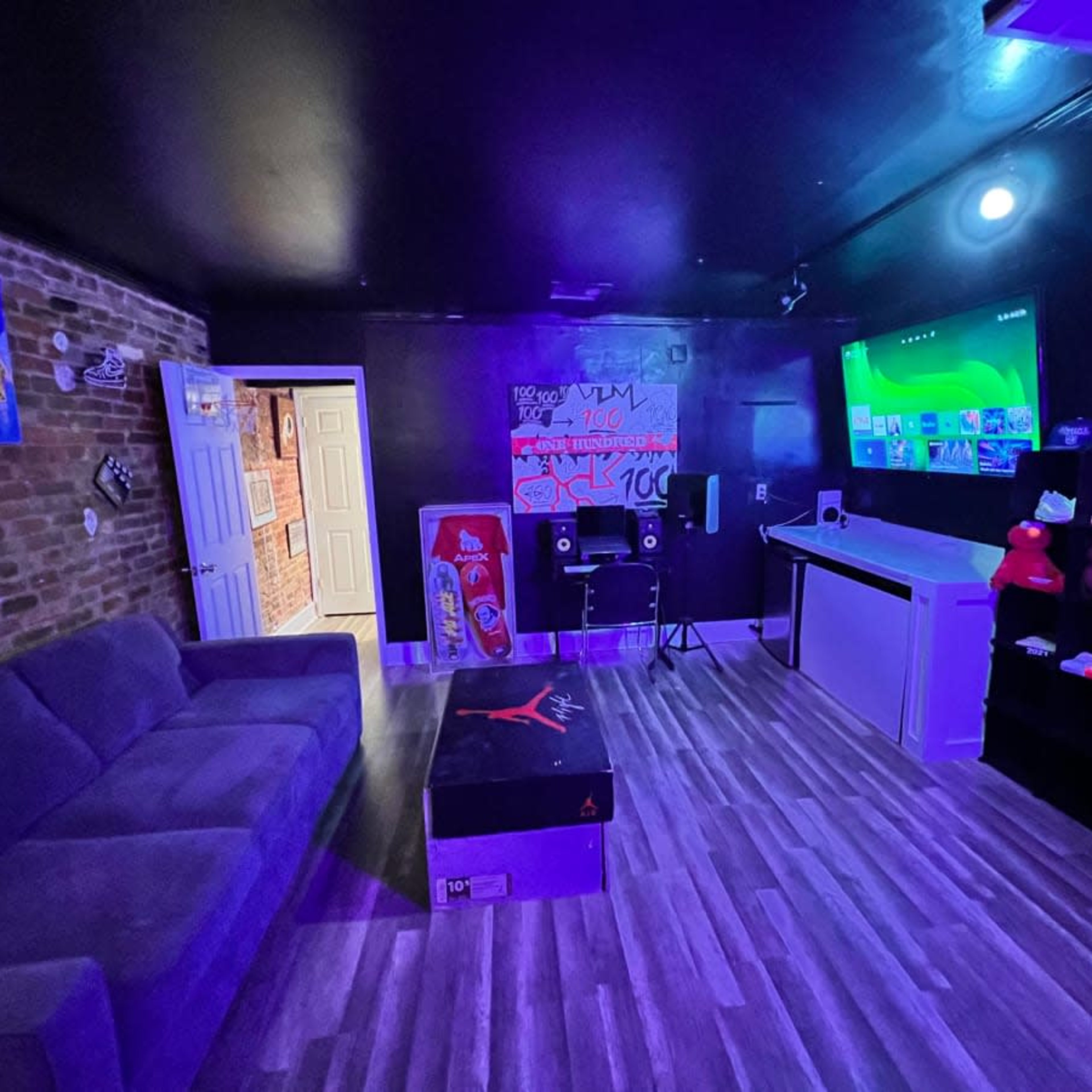 A modern game room featuring a gray sofa, a large screen on the wall, shelves with colorful shoes, and exposed brick accents.