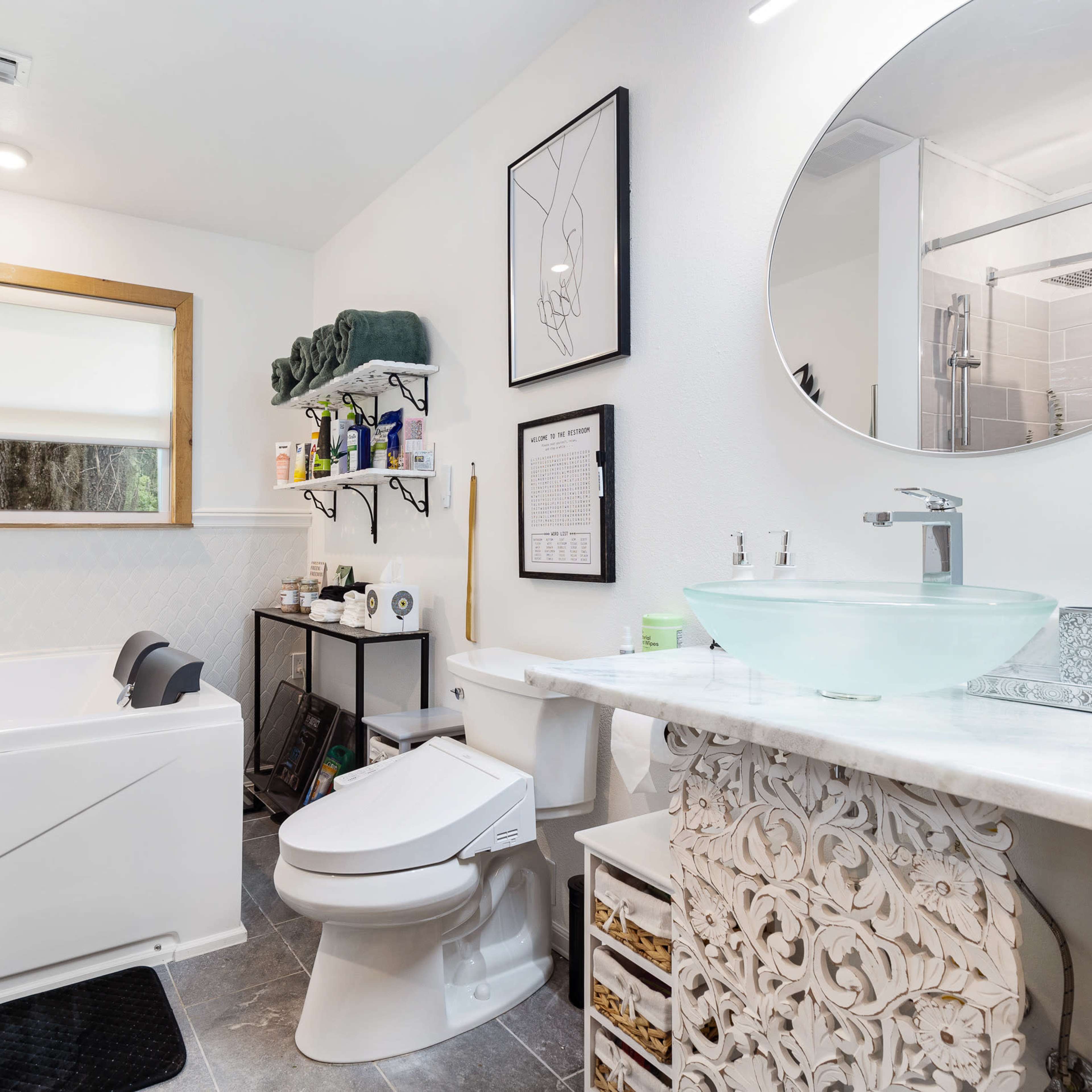 The bathroom features a shower, a bathtub with a gray chair, a toilet, a sink with a round mirror, and neatly organized shelves and storage units.