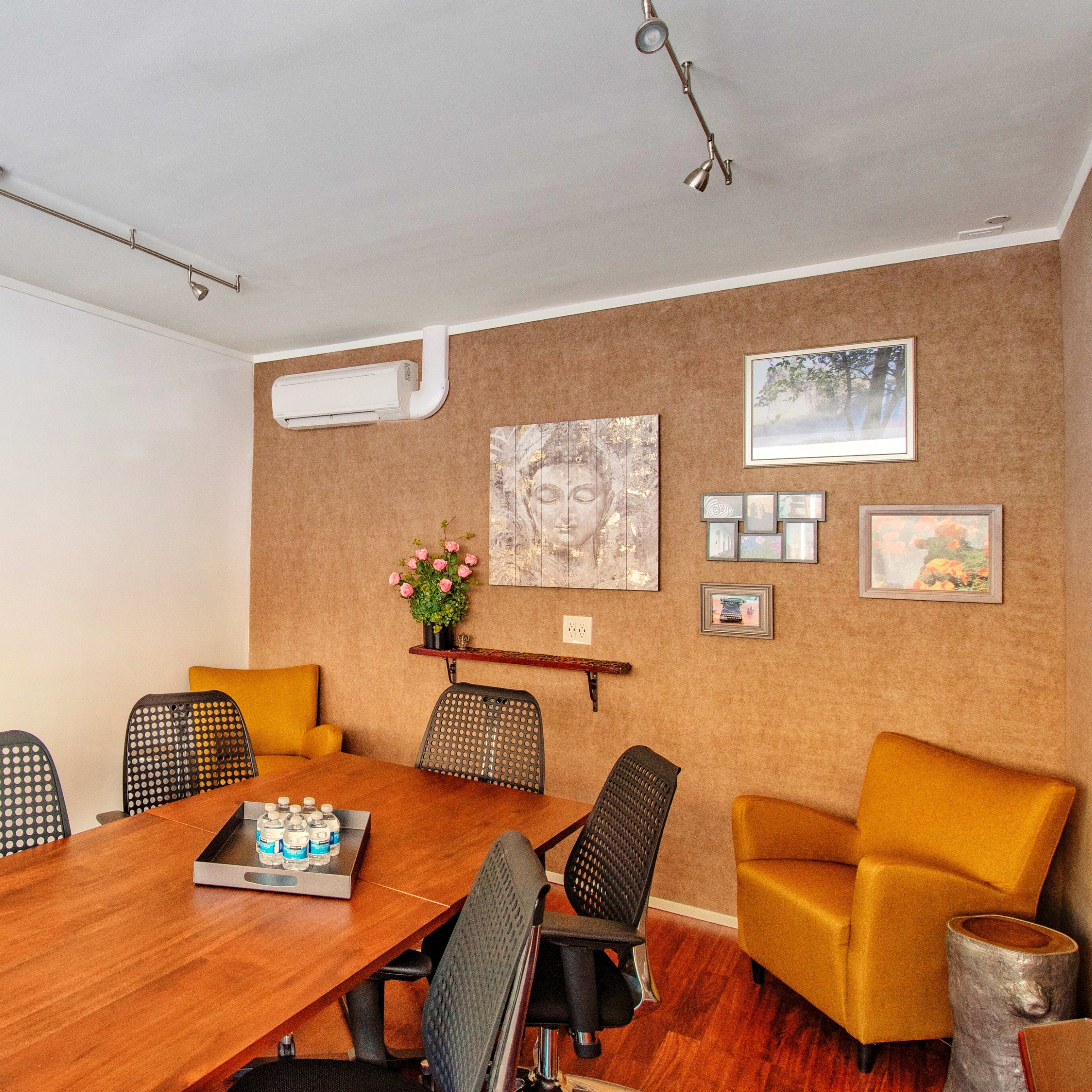 A conference room features a wooden table surrounded by black mesh chairs, with two yellow armchairs and artwork on the walls.