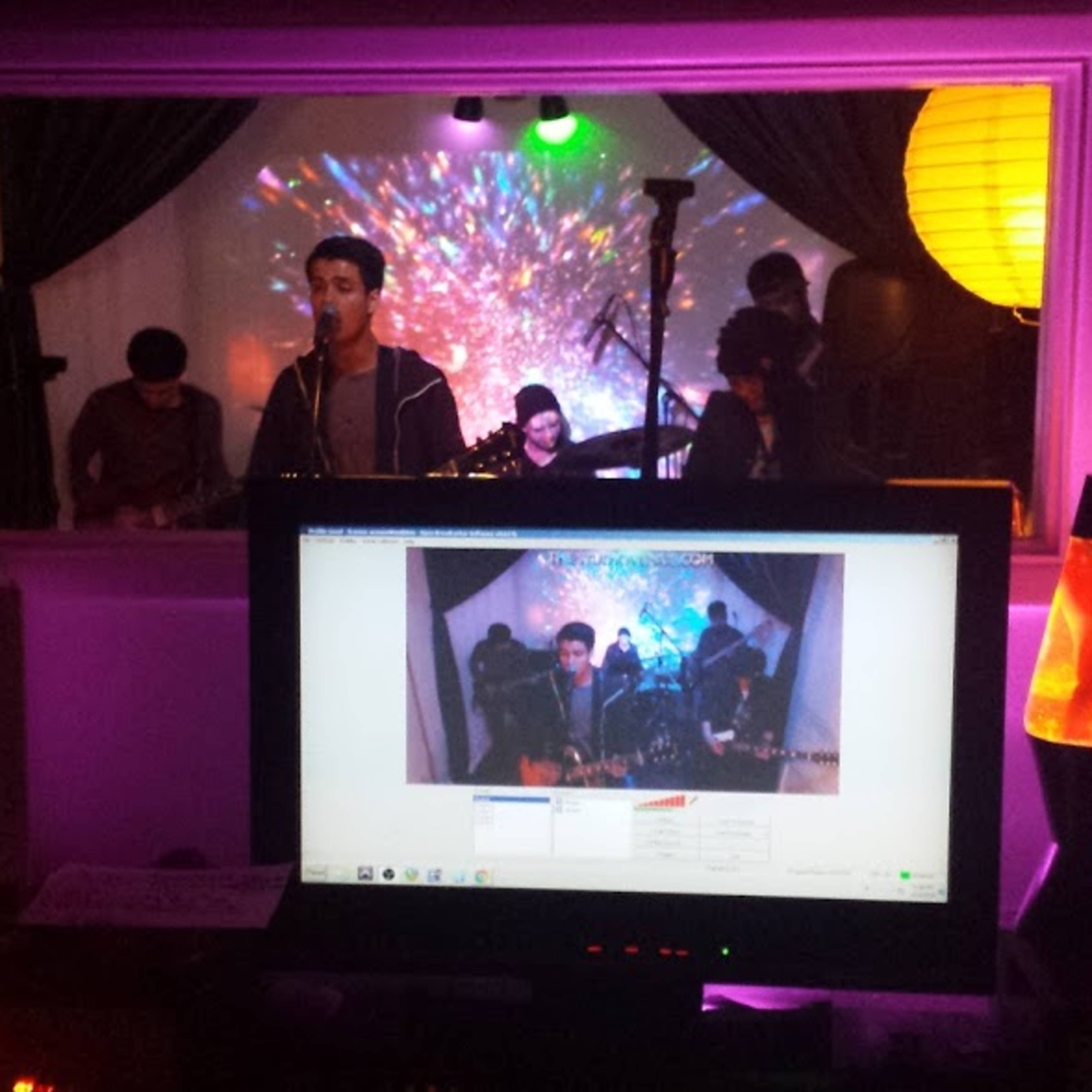 A band performs in a studio with a colorful visual backdrop, while a computer screen displays the live feed of the performance.