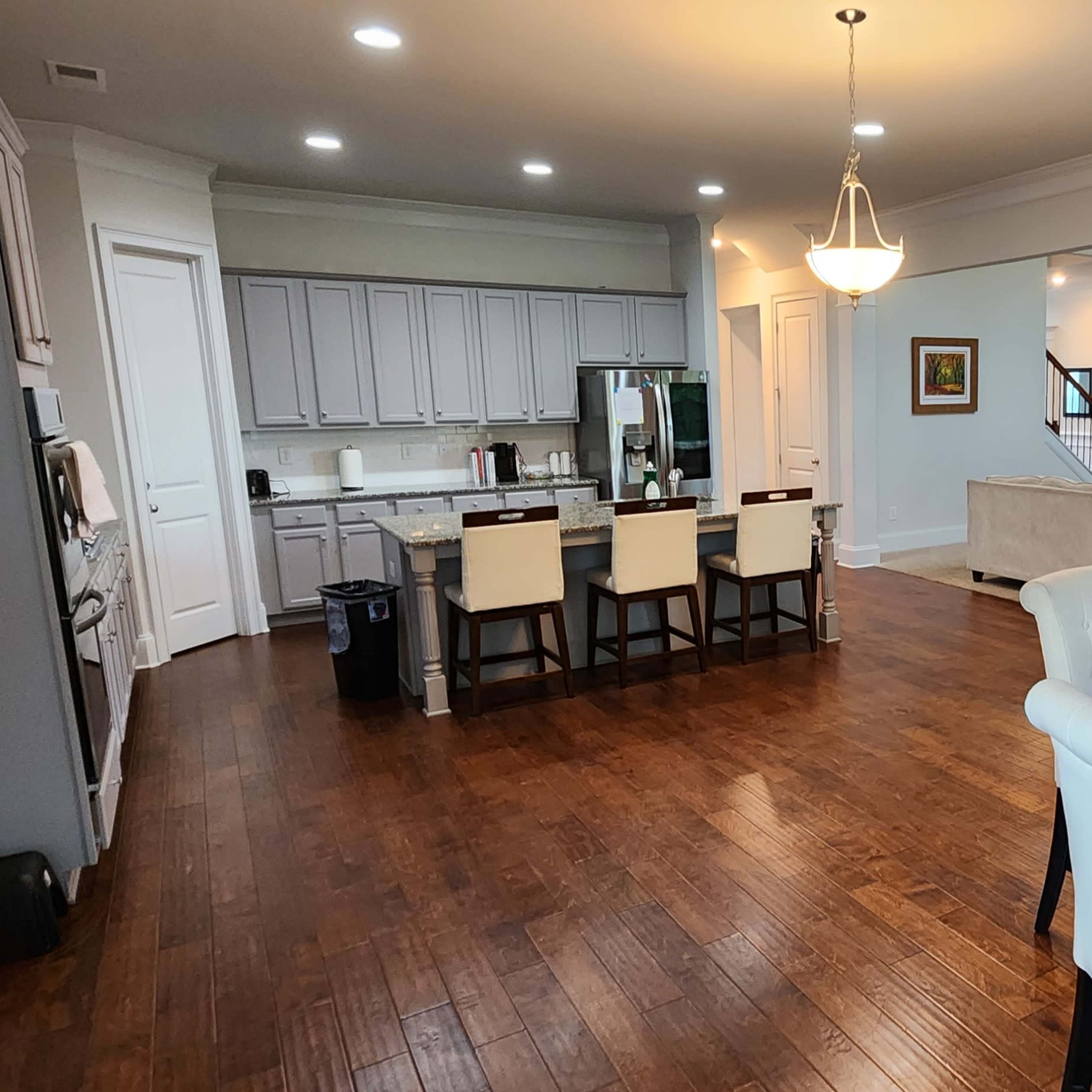 A spacious kitchen and dining area with a round wooden table and a set of bar stools near a counter, featuring light-colored cabinetry and hardwood flooring.