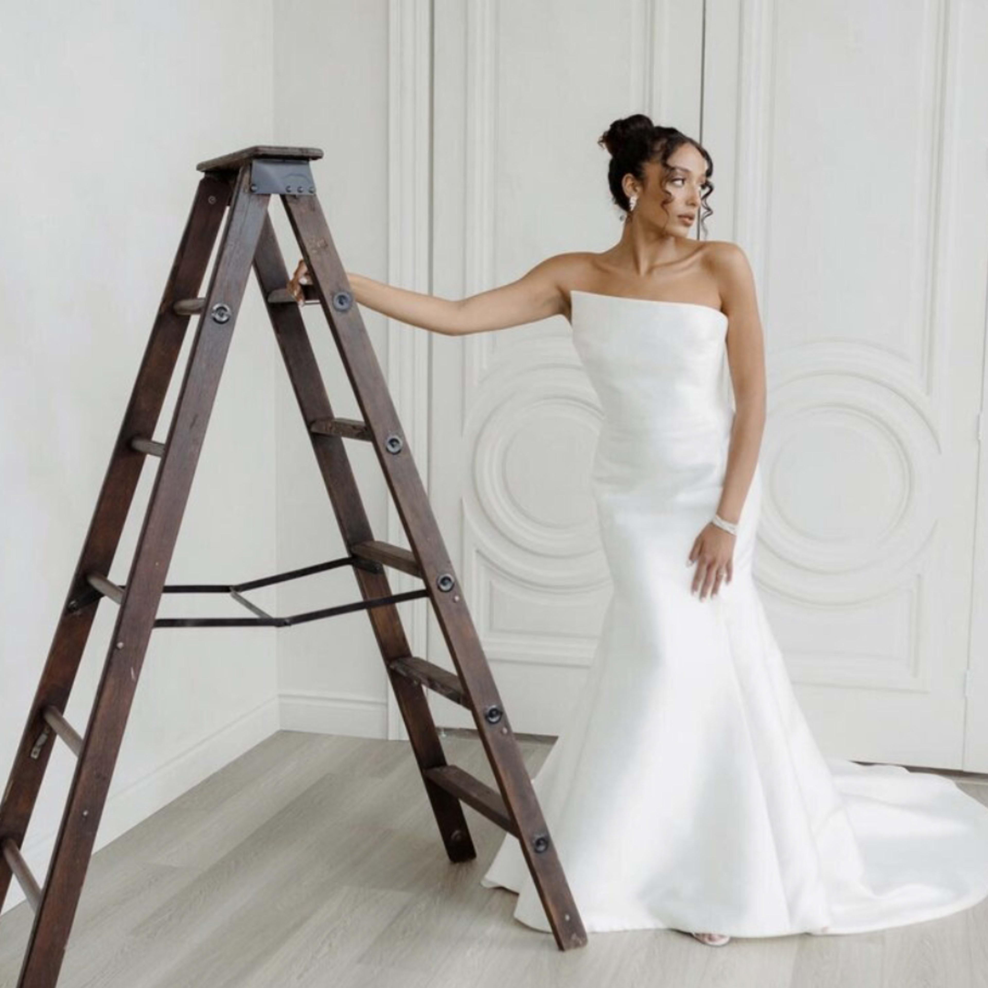 A woman in a strapless white wedding dress stands beside a wooden ladder in a room with light-colored walls and a floral backdrop.