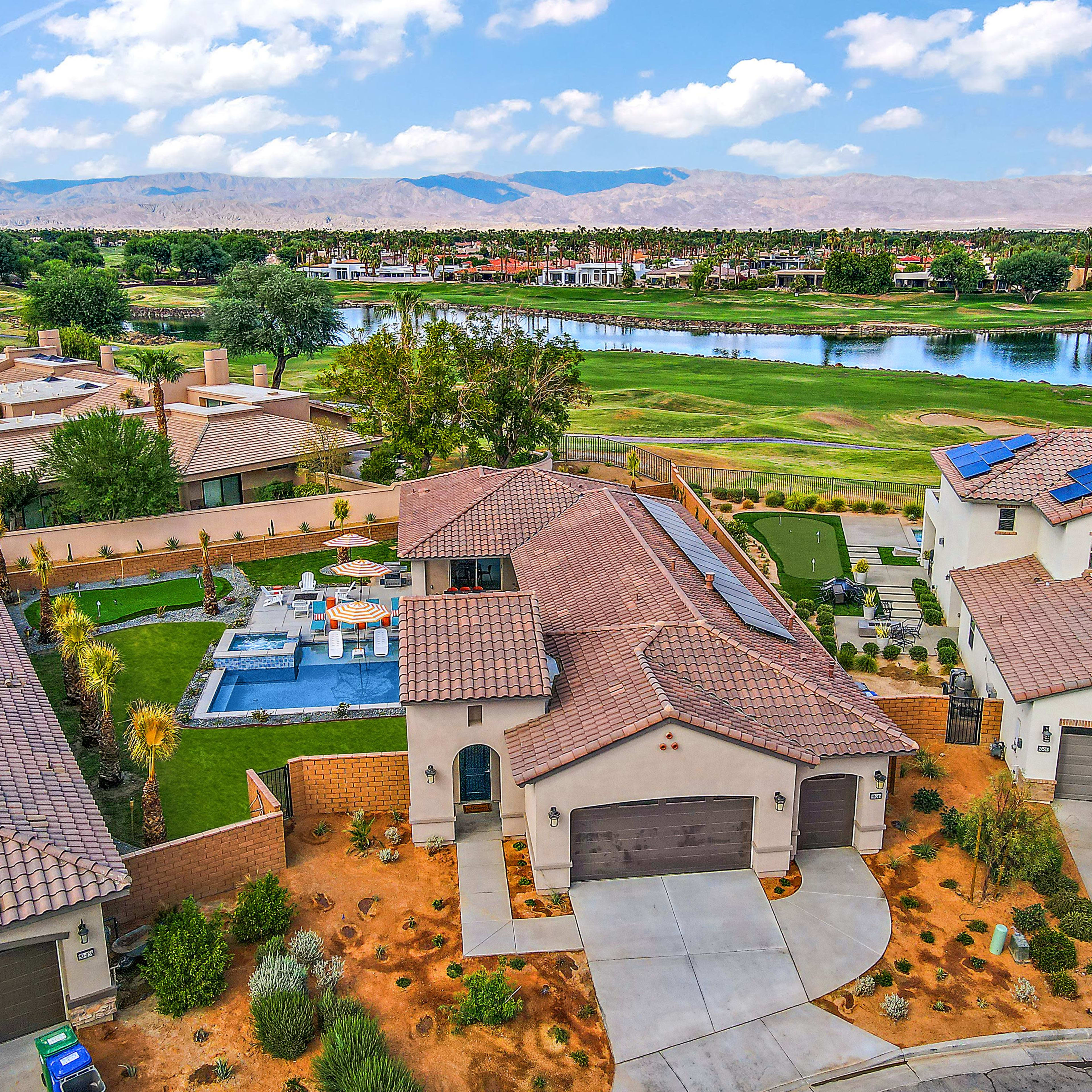A residential neighborhood featuring several houses, a swimming pool in the backyard, and a lake with mountains in the background.