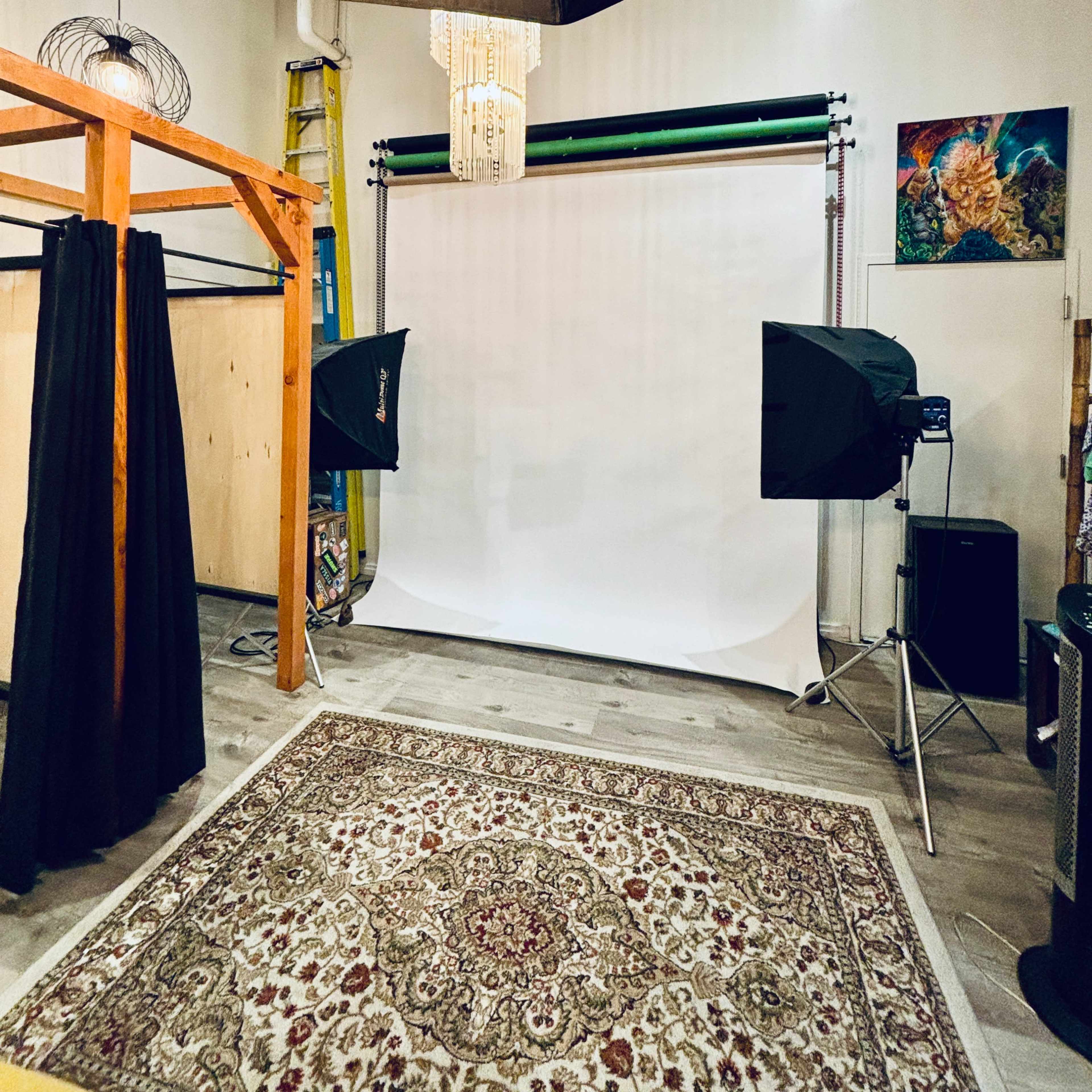 A photography studio with a white backdrop, black curtains, and lighting equipment arranged around a patterned rug.