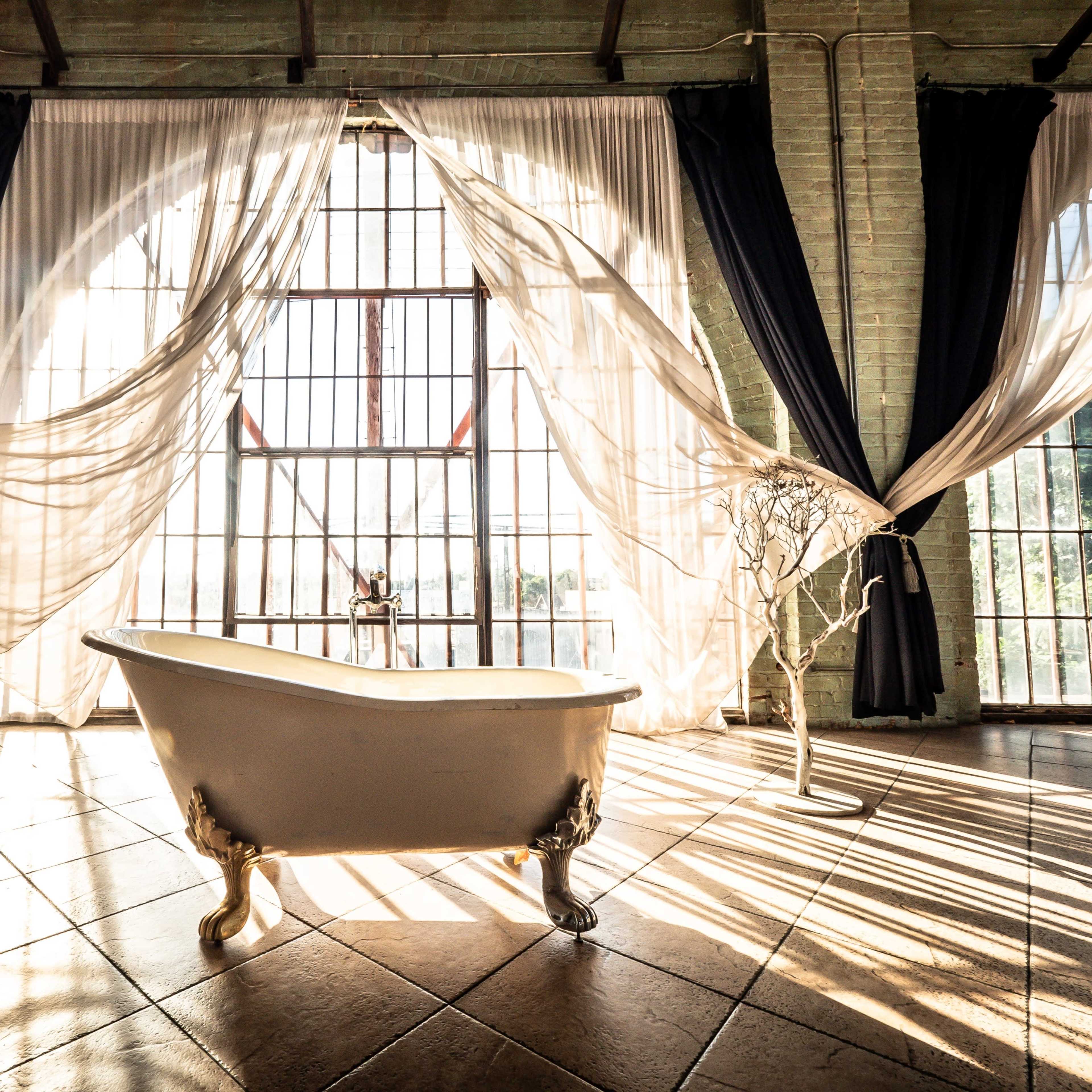 A vintage freestanding bathtub is positioned on a tiled floor beneath large windows, framed by flowing curtains and decorative trees.