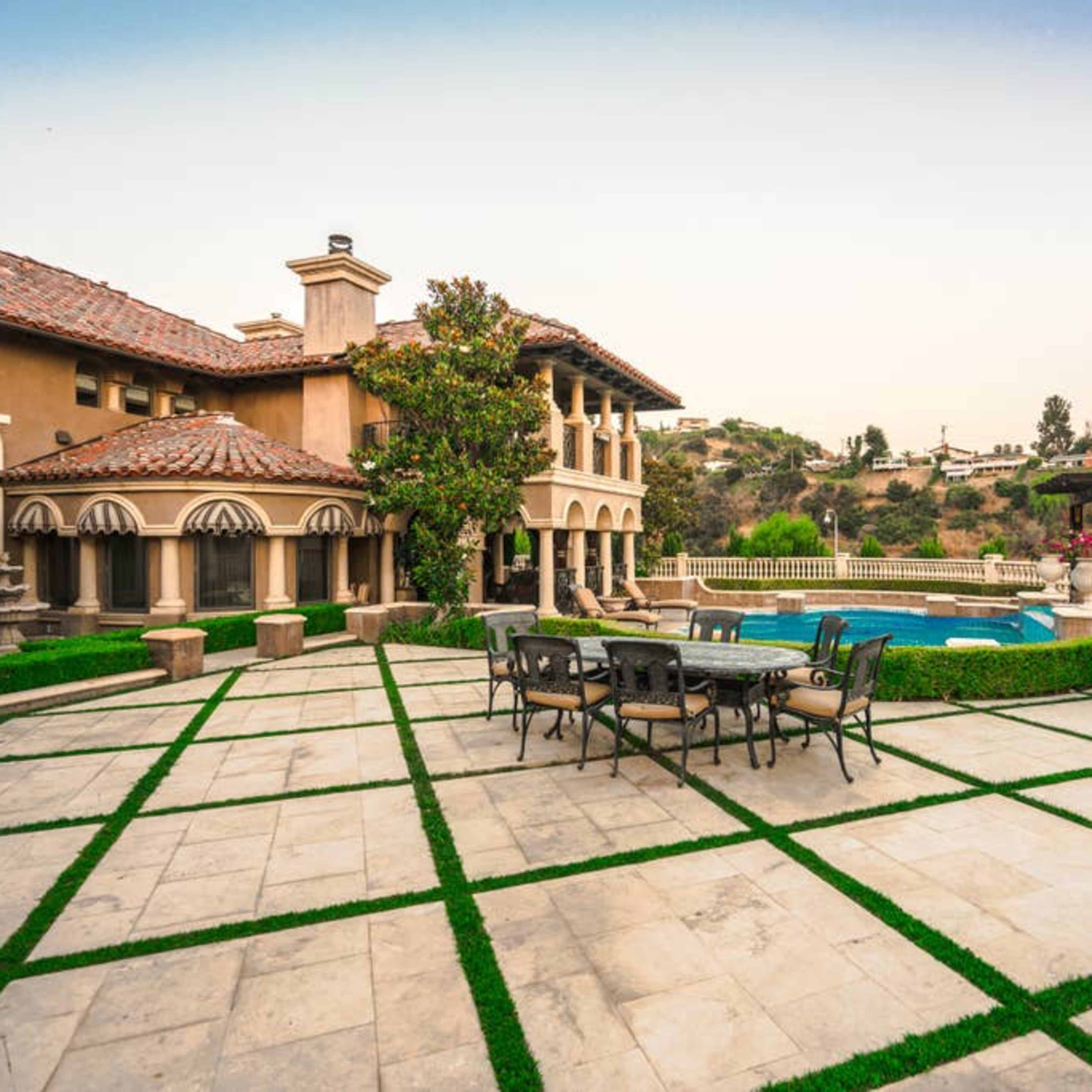 A luxury mansion with a stone patio, outdoor dining set, and a swimming pool surrounded by manicured landscaping.