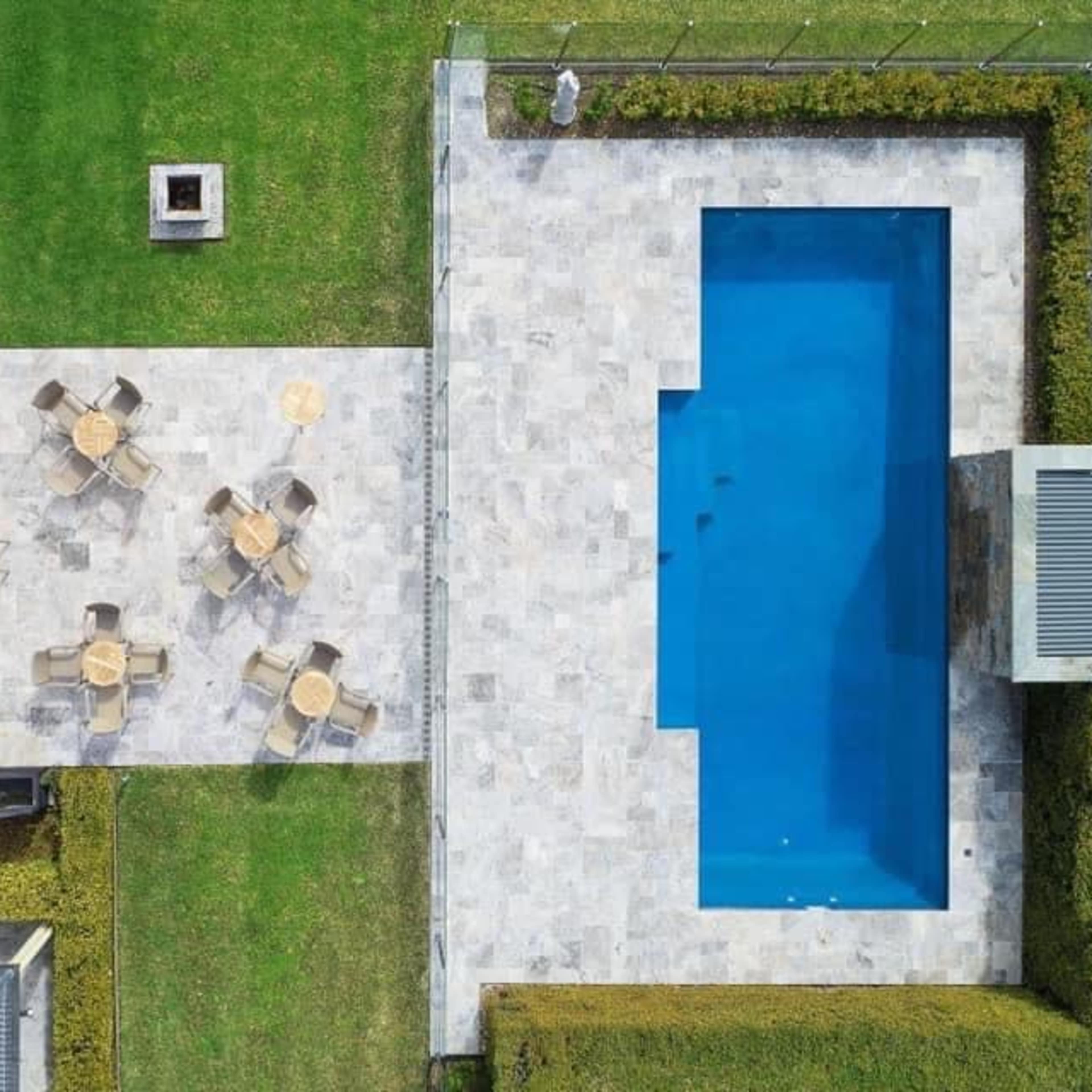 An aerial view shows a modern backyard featuring a rectangular swimming pool surrounded by a stone patio with several tables and chairs.