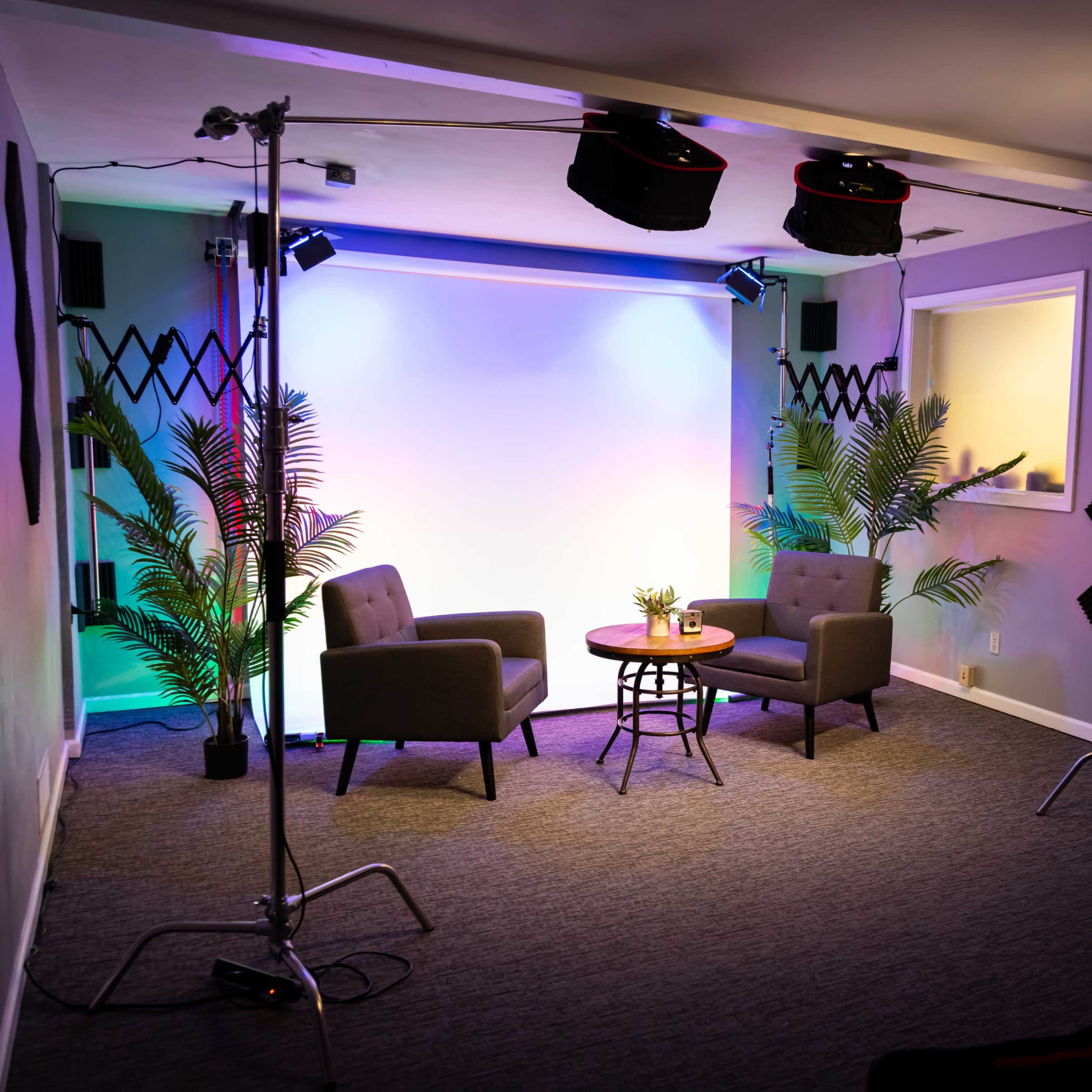 A studio setting featuring two grey chairs, a small table with a plant, and colorful lighting in the background.