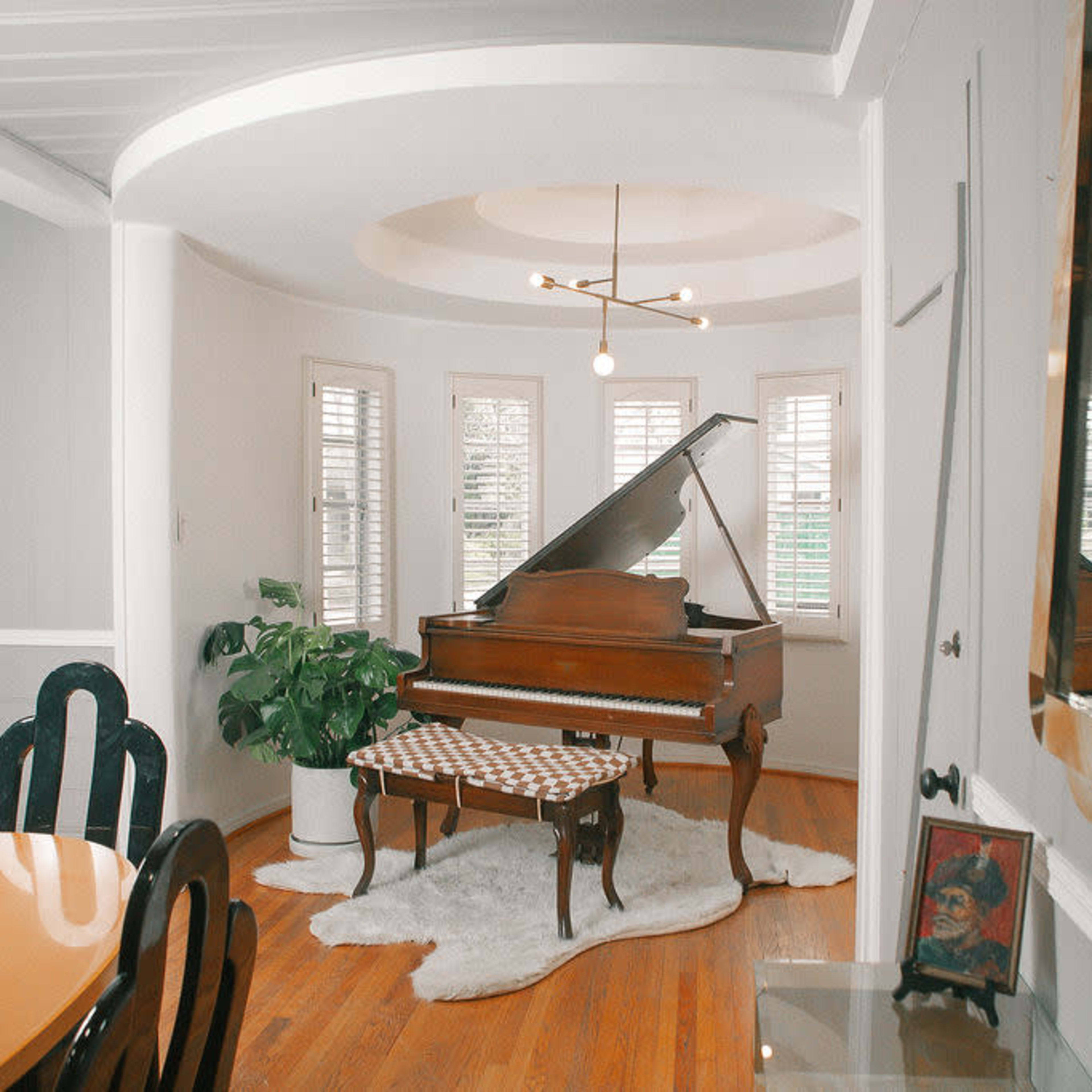 A grand piano sits in a light-filled bay window area, surrounded by potted plants and a dining table.