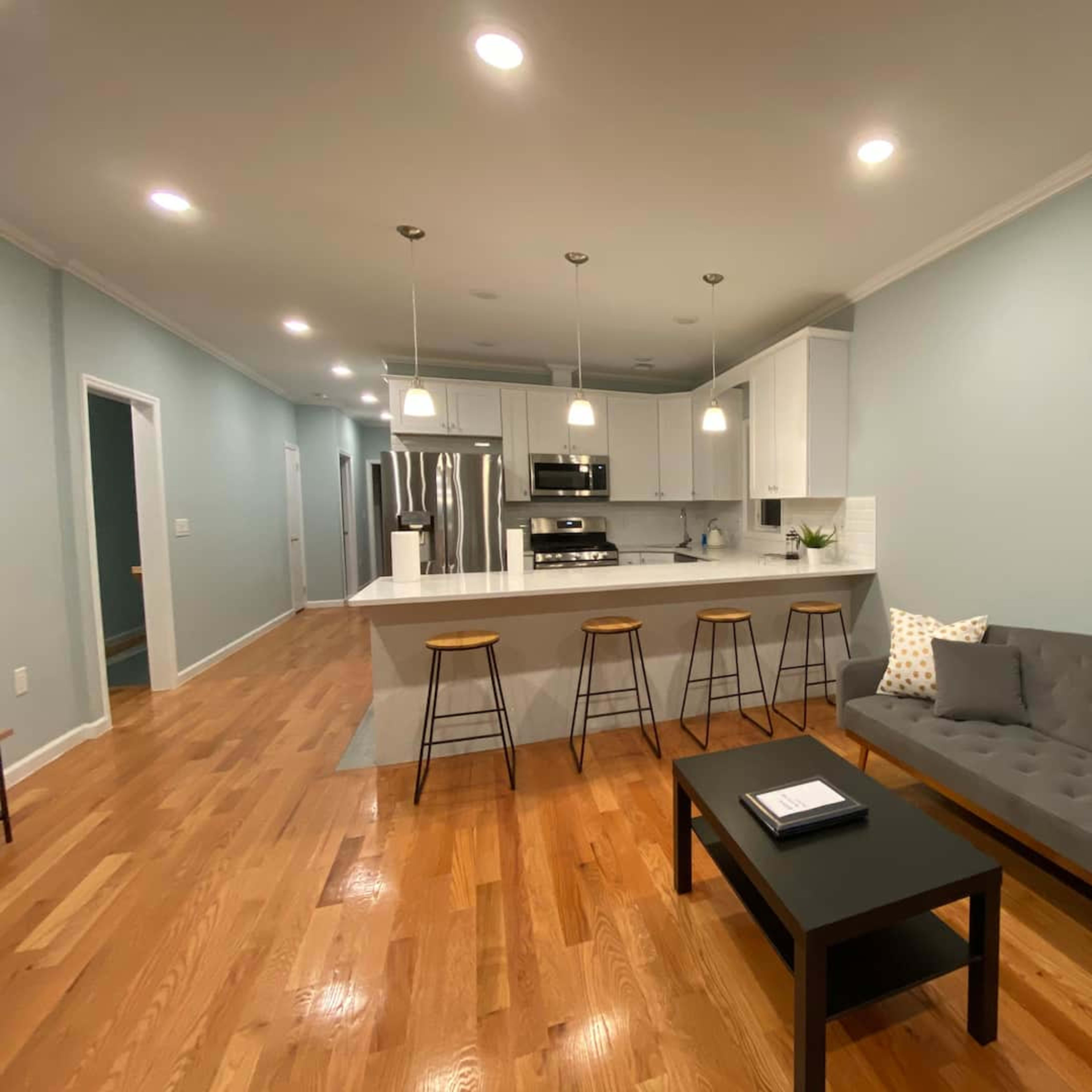 A modern living space with a gray sofa, a black coffee table, and a kitchen area featuring white cabinetry and bar stools.