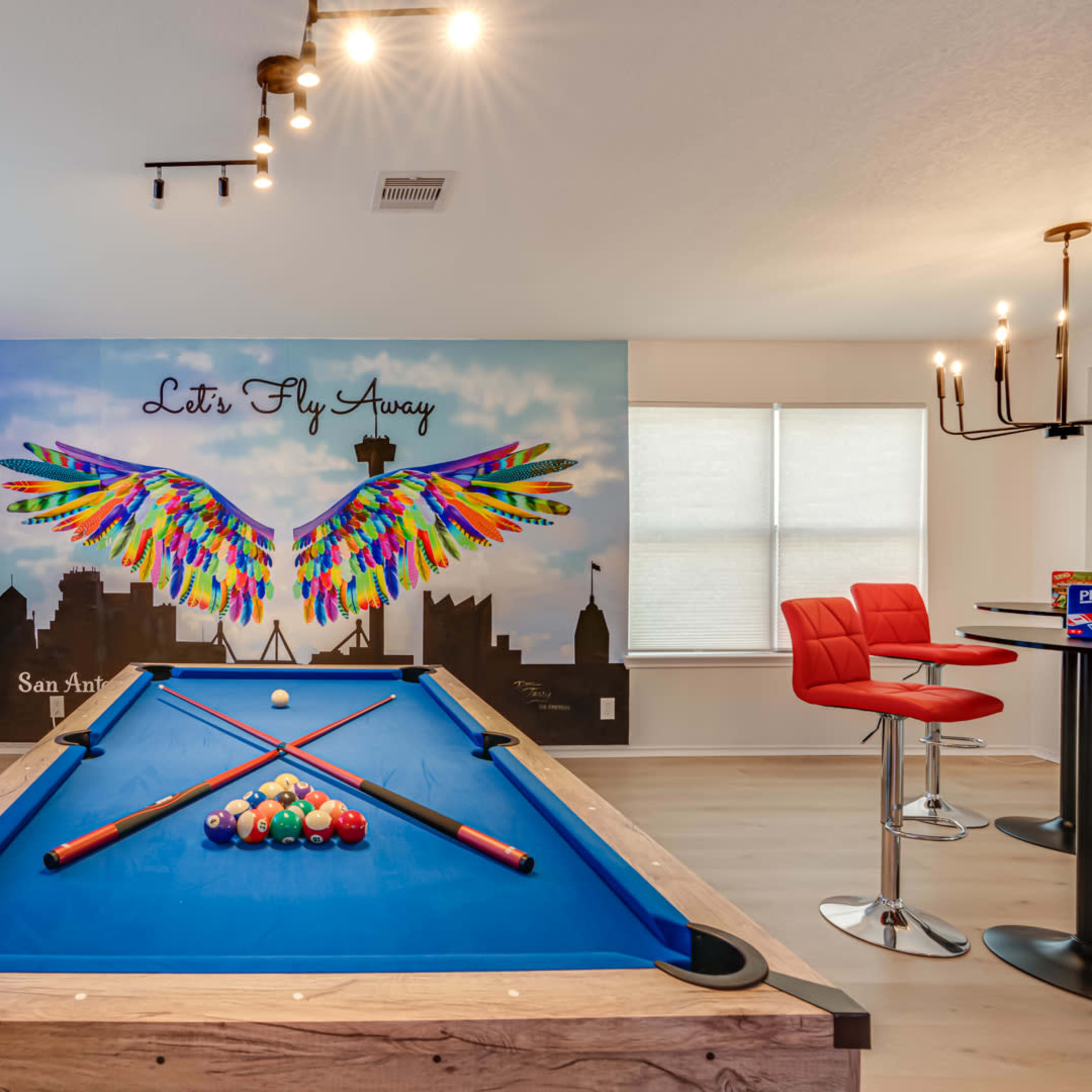 A pool table with colorful murals featuring a winged design is set against a well-lit wall with large windows and modern bar stools.
