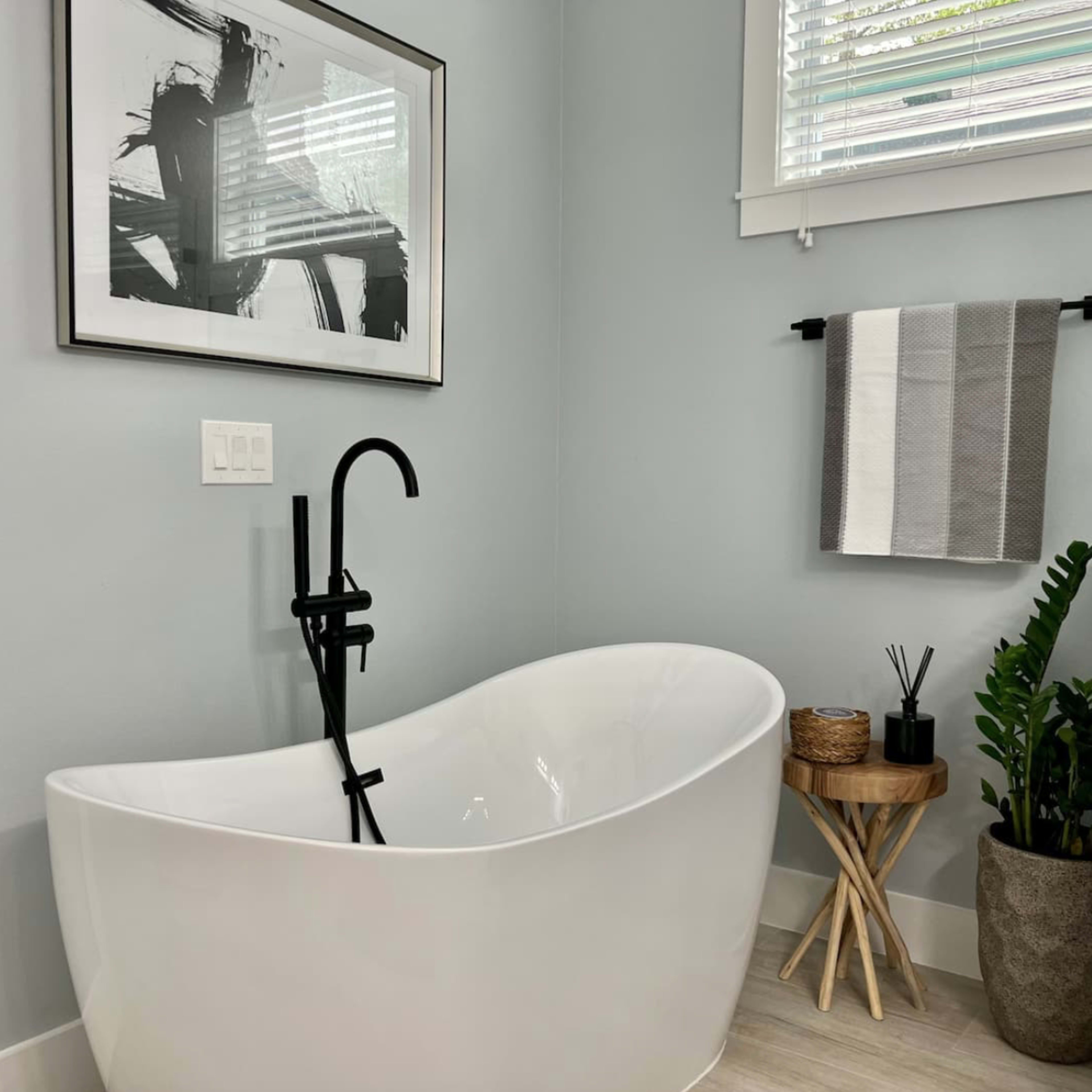 A modern bathroom features a freestanding white bathtub, a black faucet, a towel rack with gray towels, and a potted plant beside a small wooden stool.
