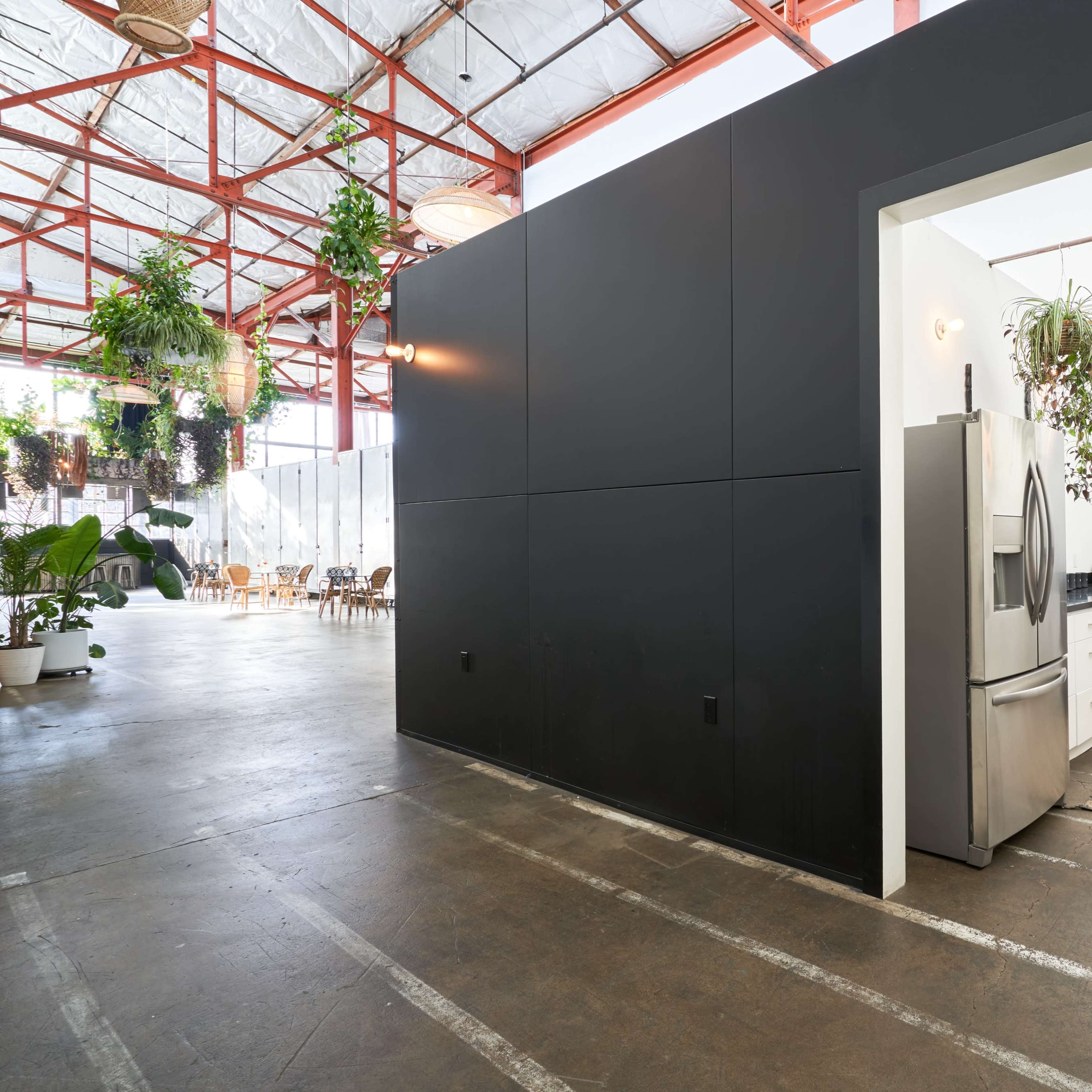 A modern kitchen area with stainless steel appliances, sleek white cabinets, and hanging plants, adjacent to a spacious dining area under a high ceiling.