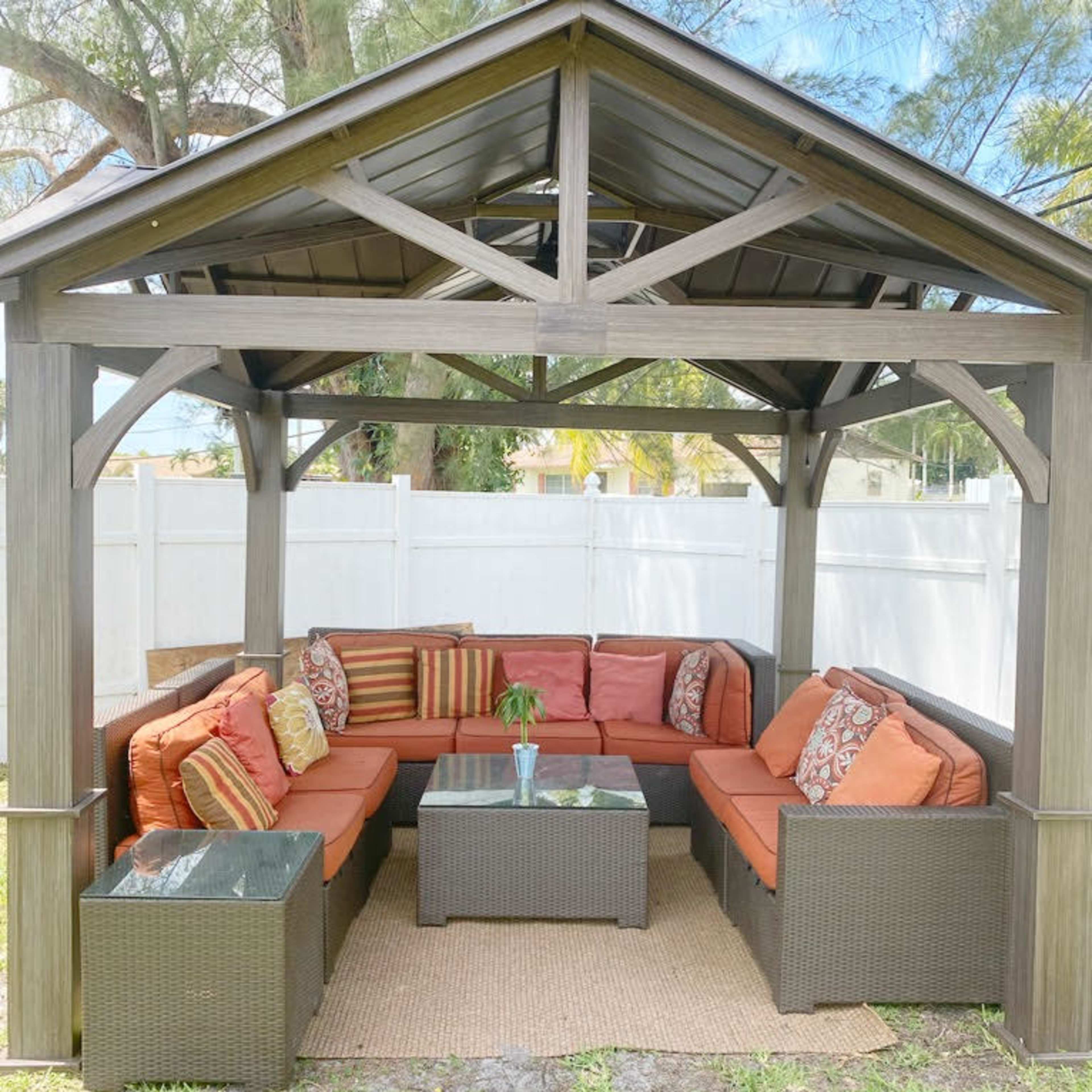 A covered outdoor seating area furnished with a sectional sofa, decorative cushions, and a coffee table, situated on a lawn next to a white fence.