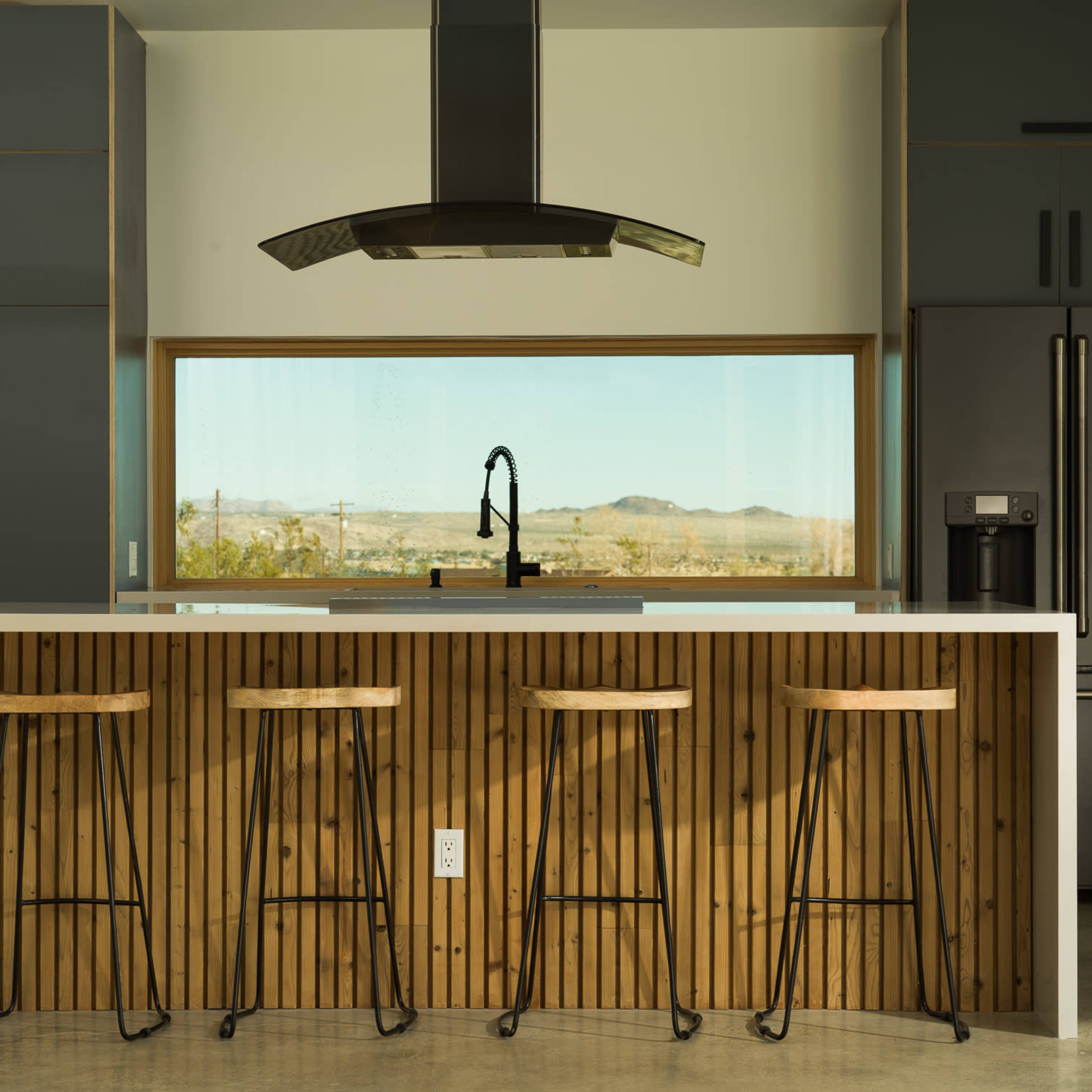 A modern kitchen features a central island with four bar stools, a sleek hood above the cooktop, and large windows offering a view of the landscape outside.
