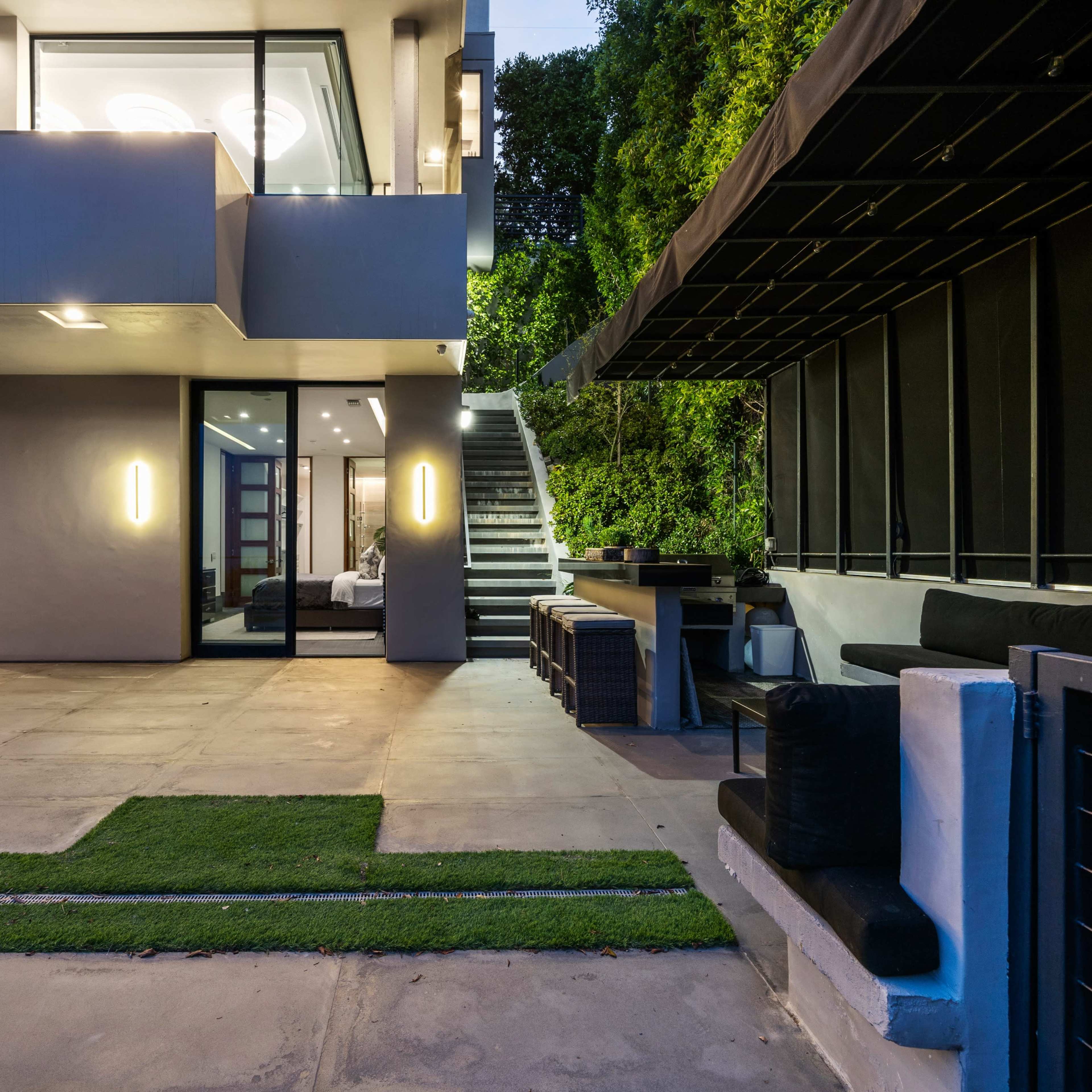 A modern exterior of a house with a patio area, green grass patches, and an outdoor staircase leading to a second floor.