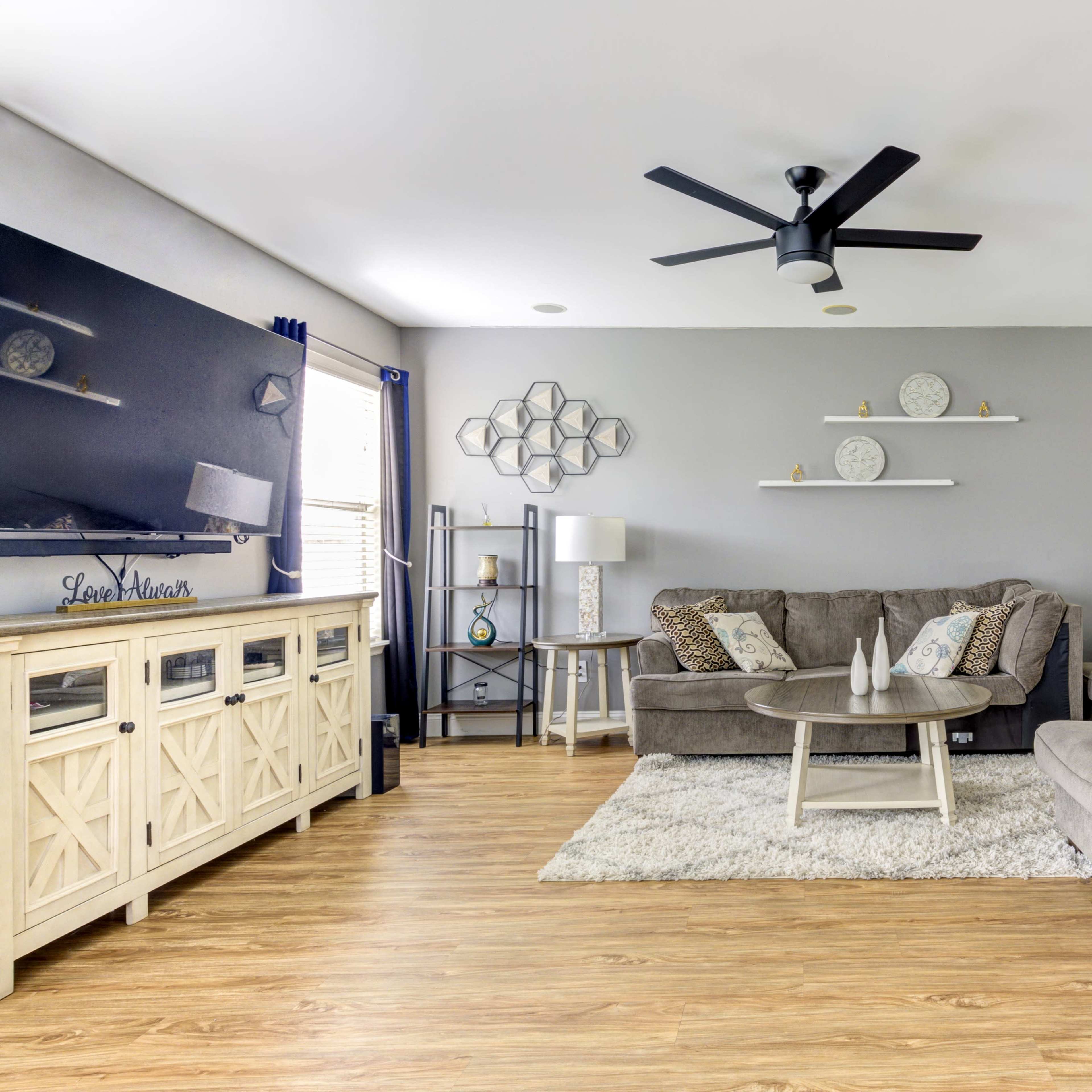 The living room features a gray sofa and loveseat arranged around a circular coffee table, with a television and decorative shelves on the wall.