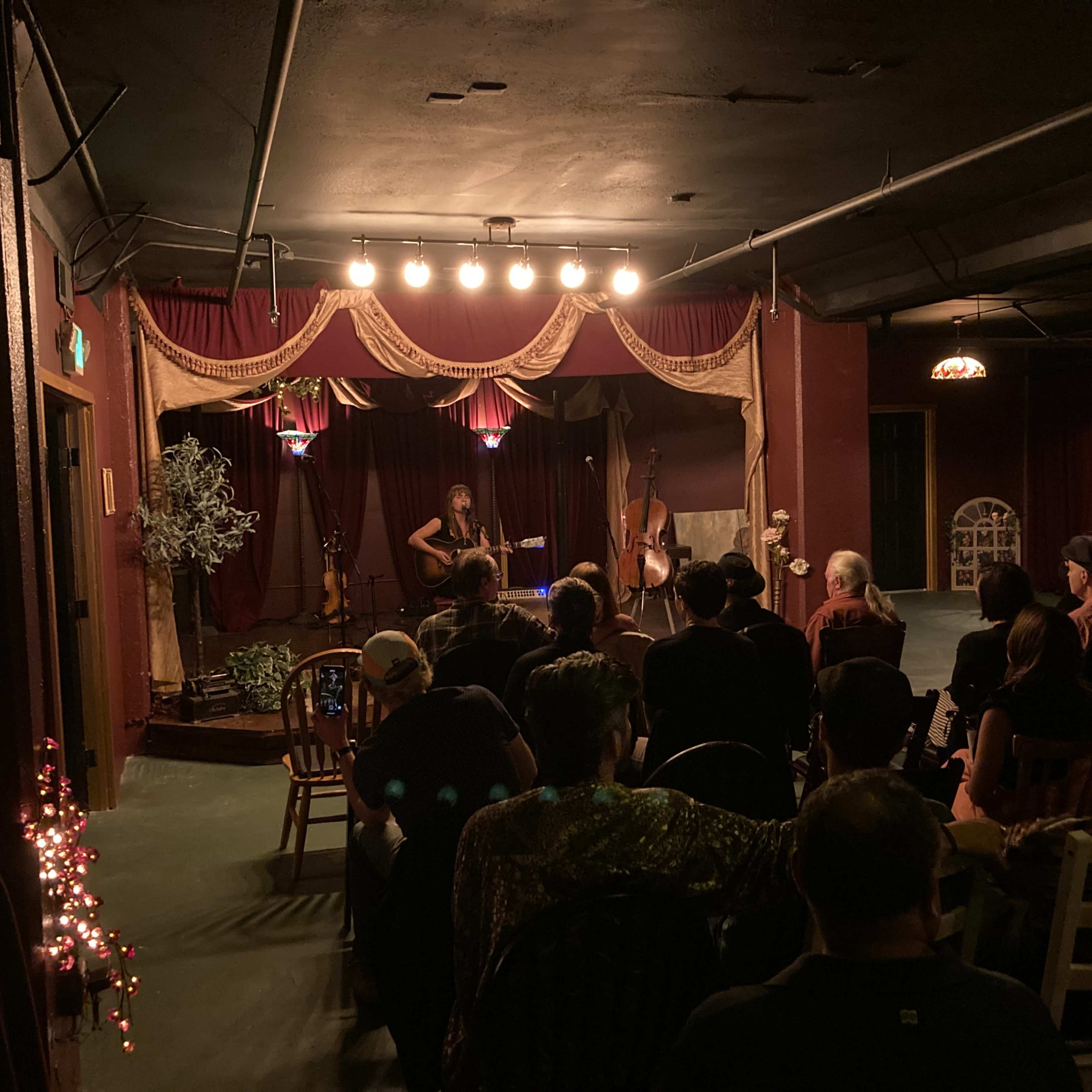 A musician performs with a guitar on stage in a dimly lit venue while an audience sits at tables.