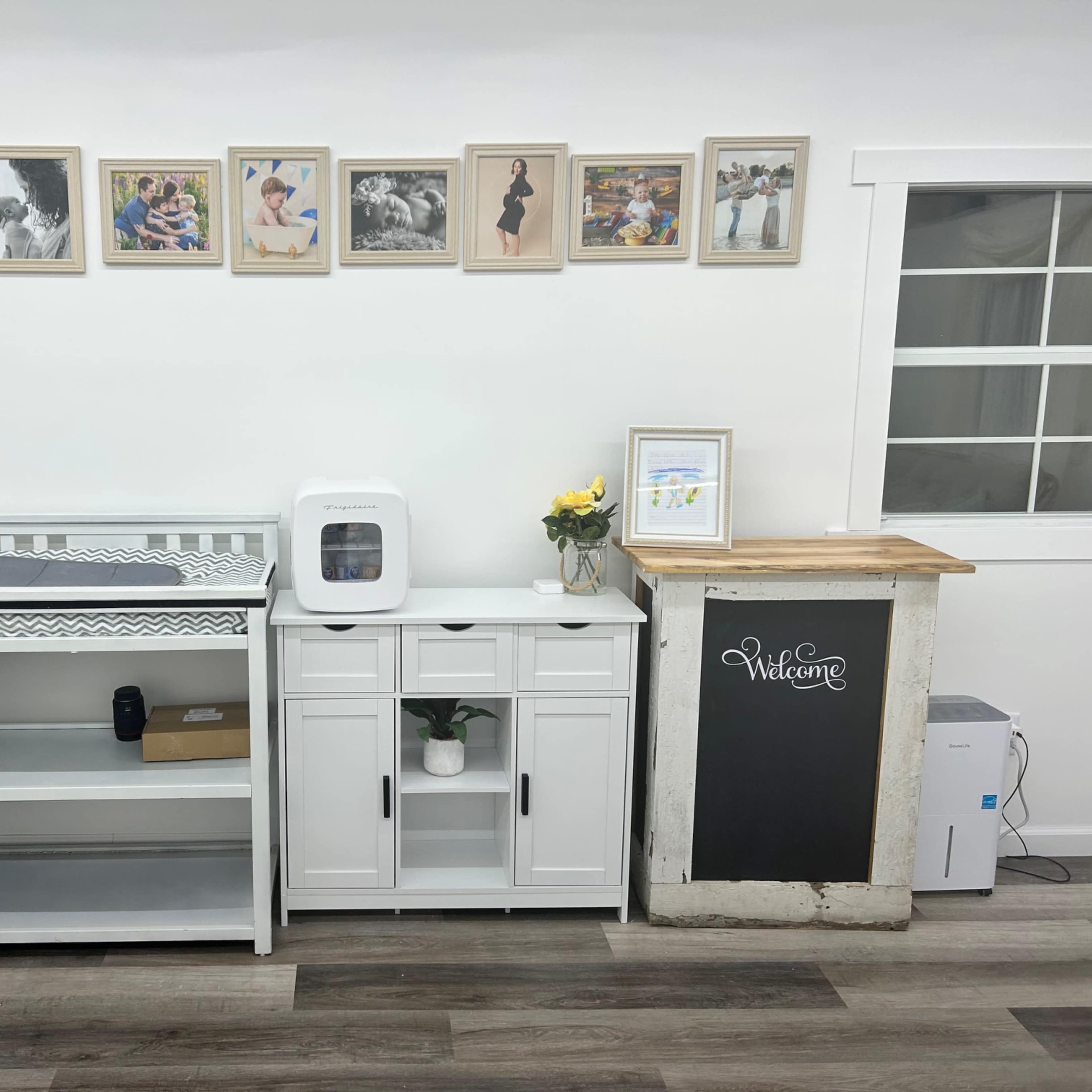 A clean white wall with a series of framed photos above a wooden reception desk, a small cabinet, and a changing table, all arranged in a well-organized space.
