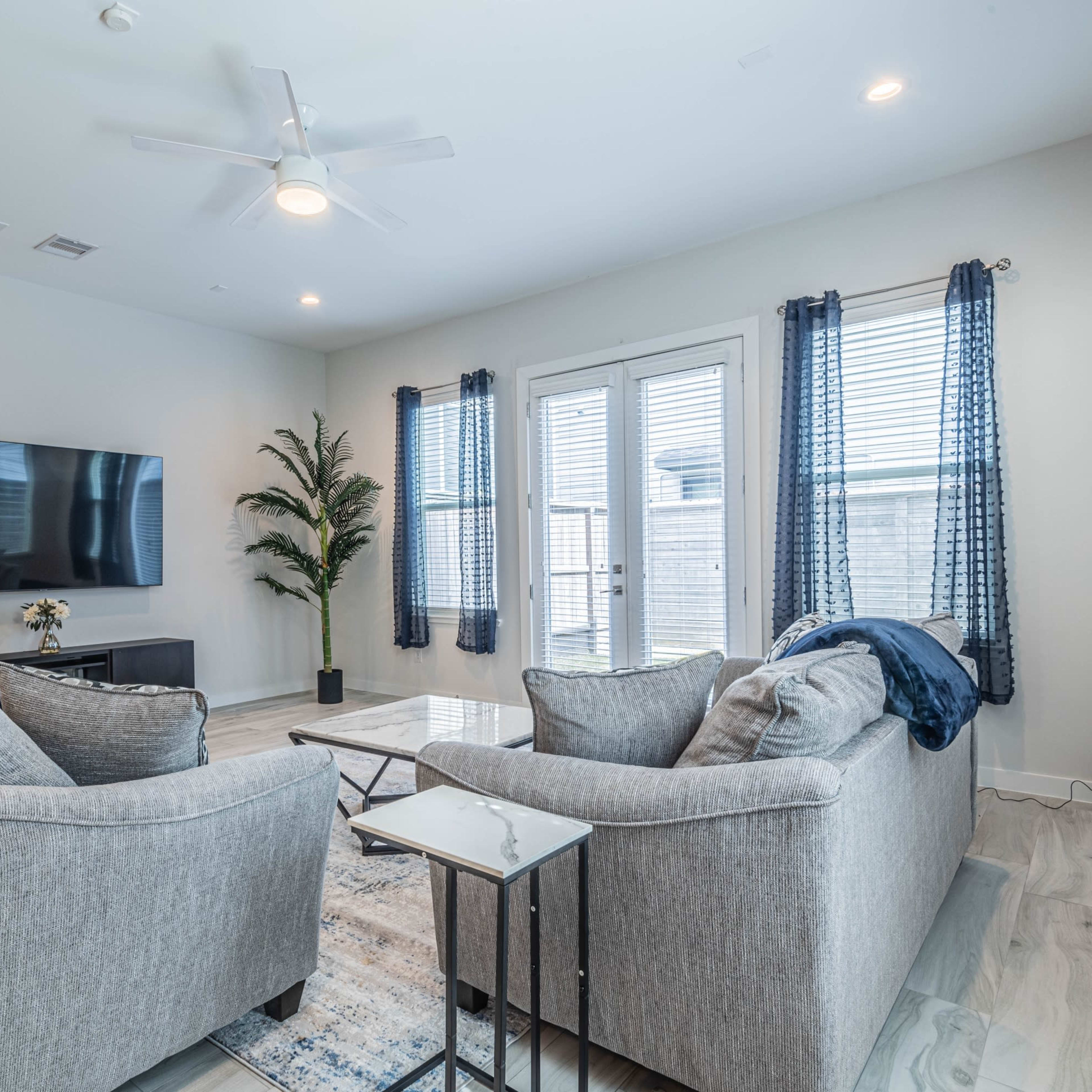 A modern living room features two gray sofas facing a coffee table, with a television mounted on the wall and large windows framed by blue curtains.