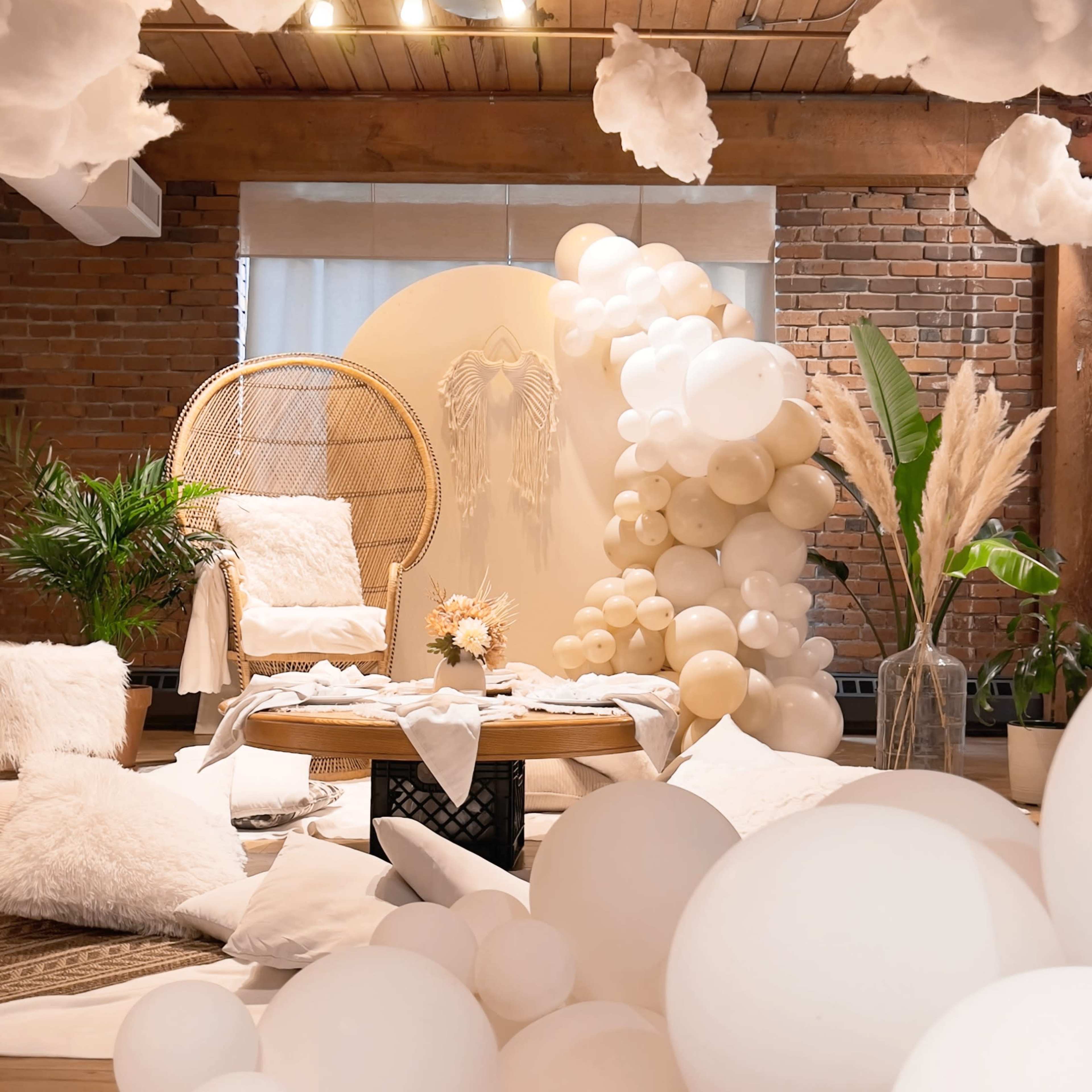 A decorated interior space features a cozy seating area with a rattan chair, a backdrop of balloons, and white cloud-like decorations hanging from the ceiling.