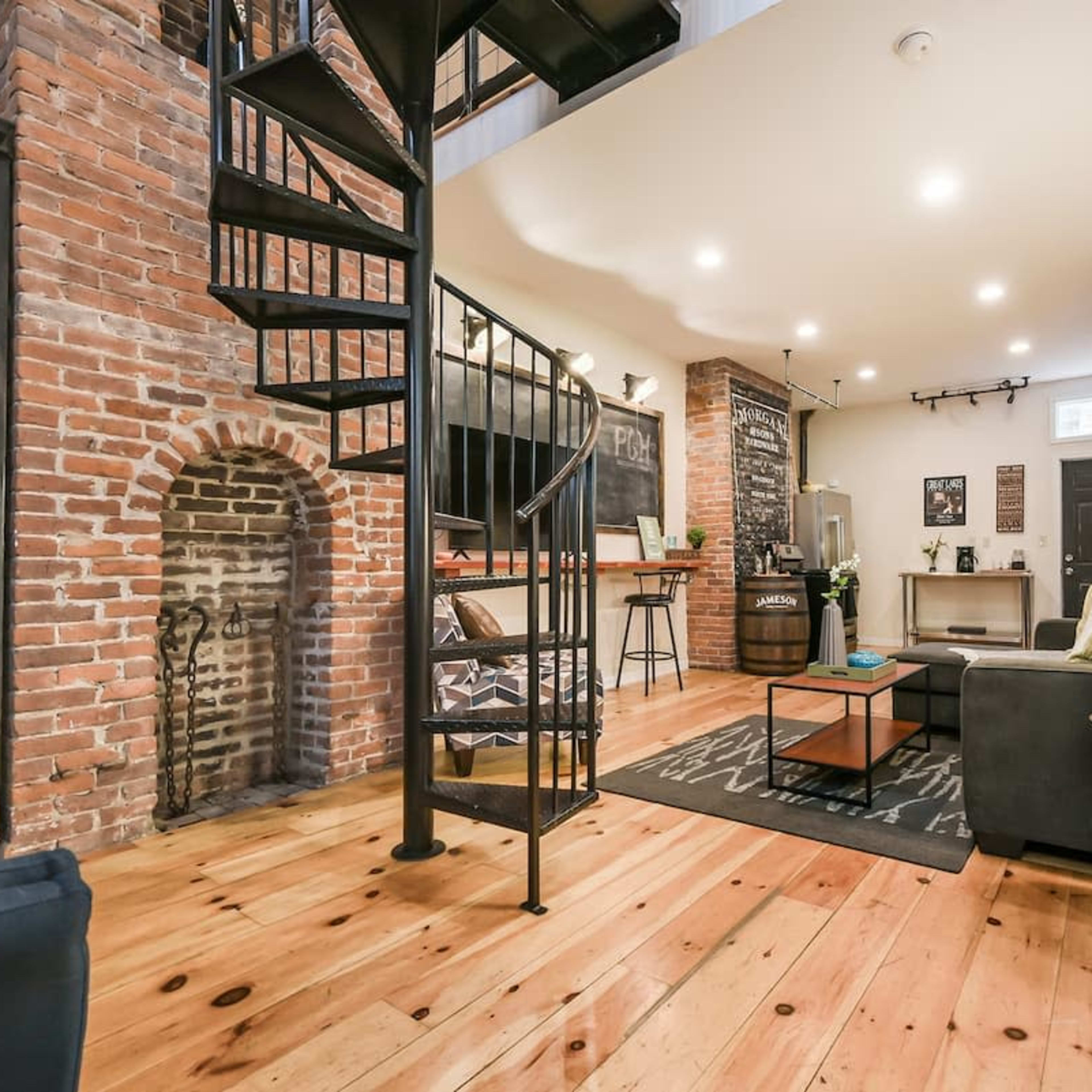 A cozy living space featuring exposed brick walls, a spiral staircase, and wooden flooring, with a seating area and a small bar.