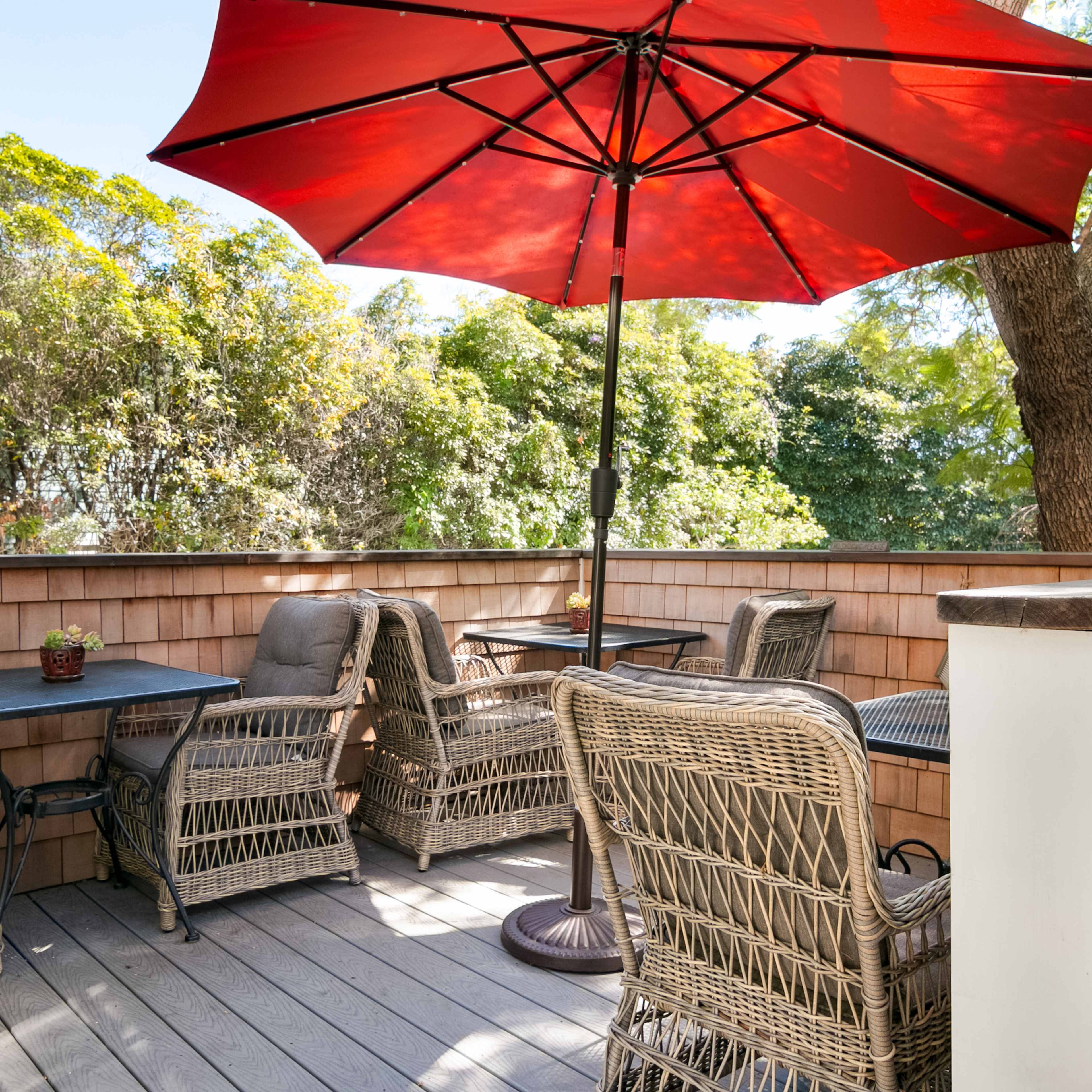 A patio with a red umbrella shades wicker furniture arranged around small tables, surrounded by greenery.