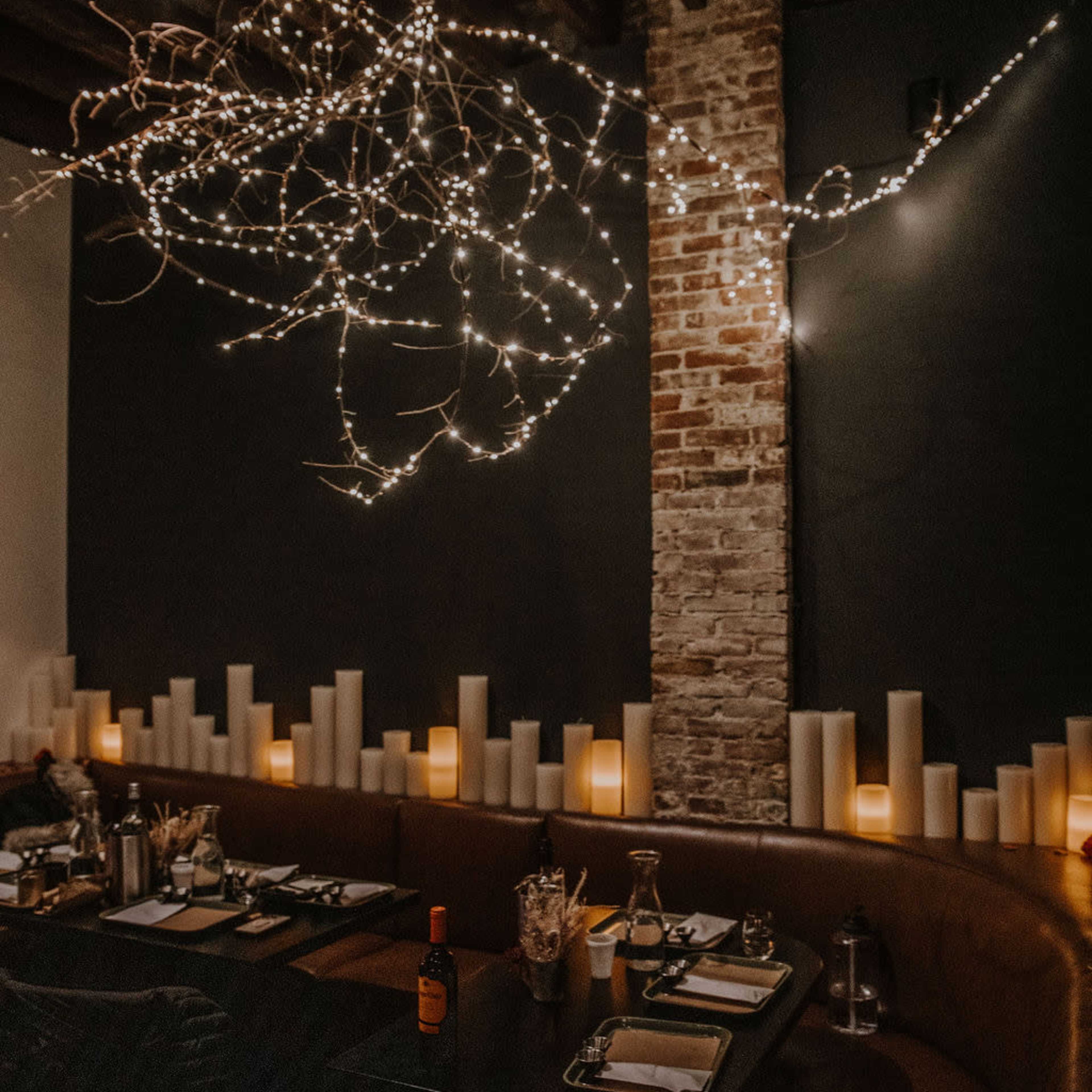 The scene features a dimly lit restaurant with string lights arranged above a table set with utensils and decor, surrounded by tall candle-like installations.