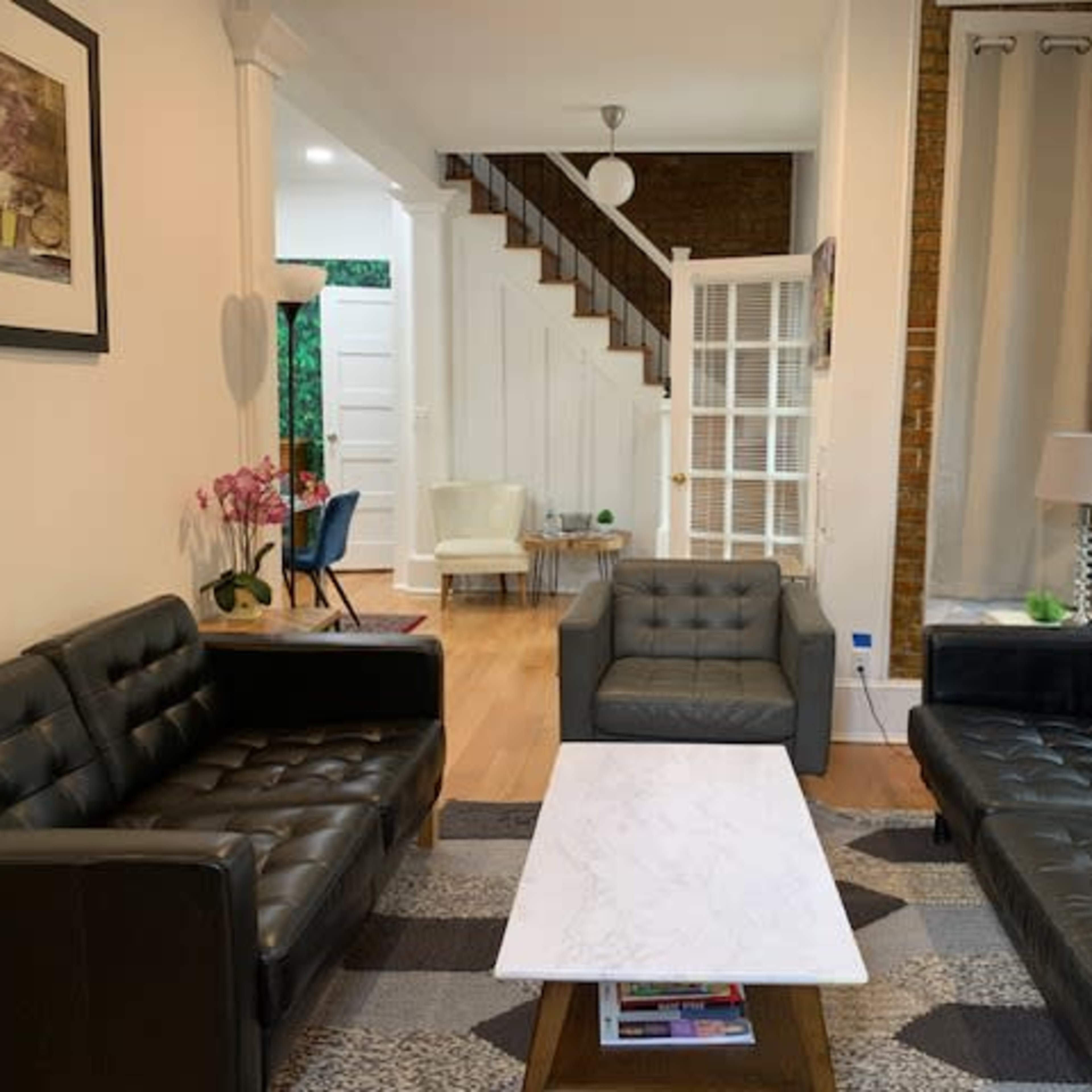 A living room with two black leather sofas facing a marble coffee table, featuring a staircase in the background and a light-filled space with decorative elements.