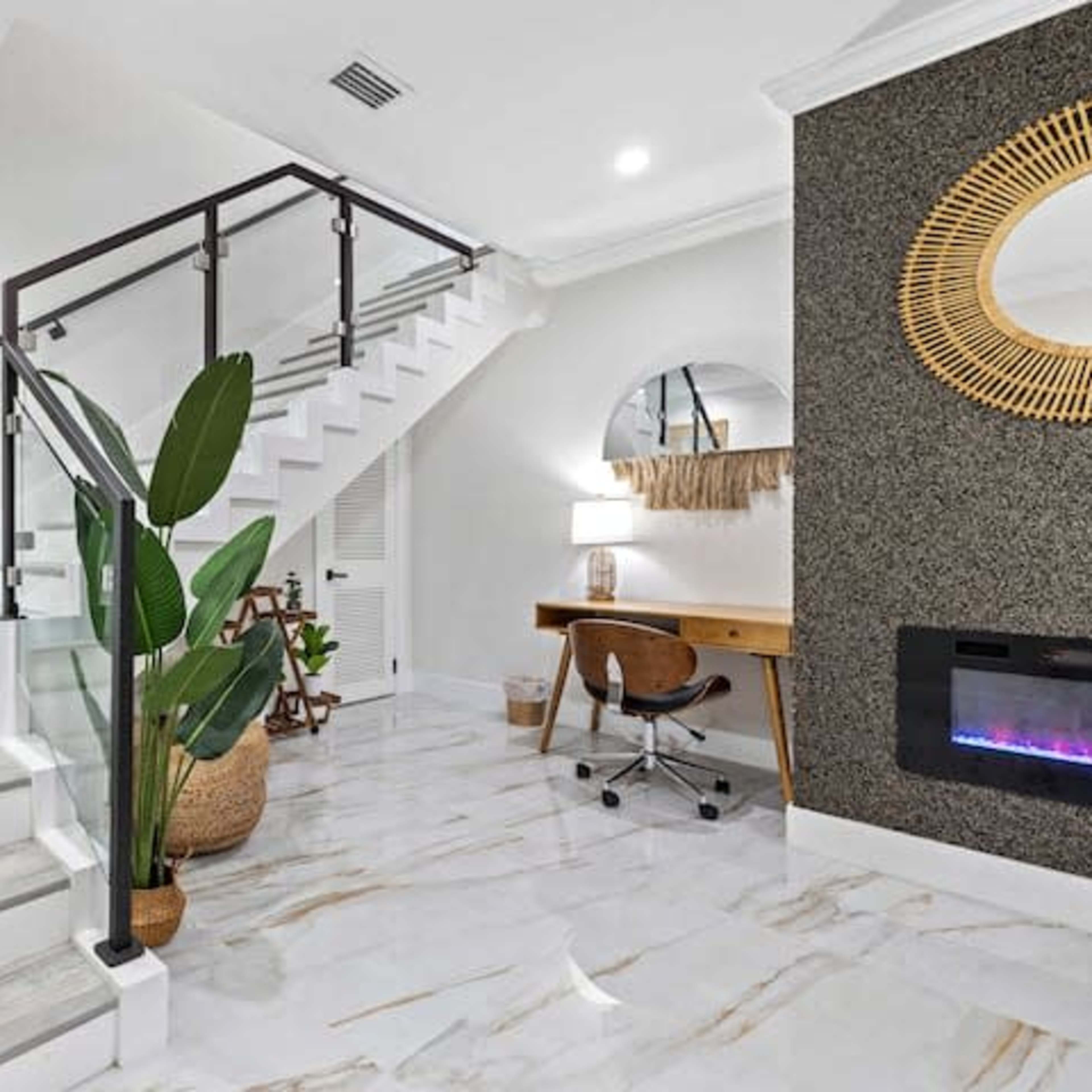 A modern staircase with glass railings, surrounded by decorative plants and a small desk area, all set in a room with marble flooring.
