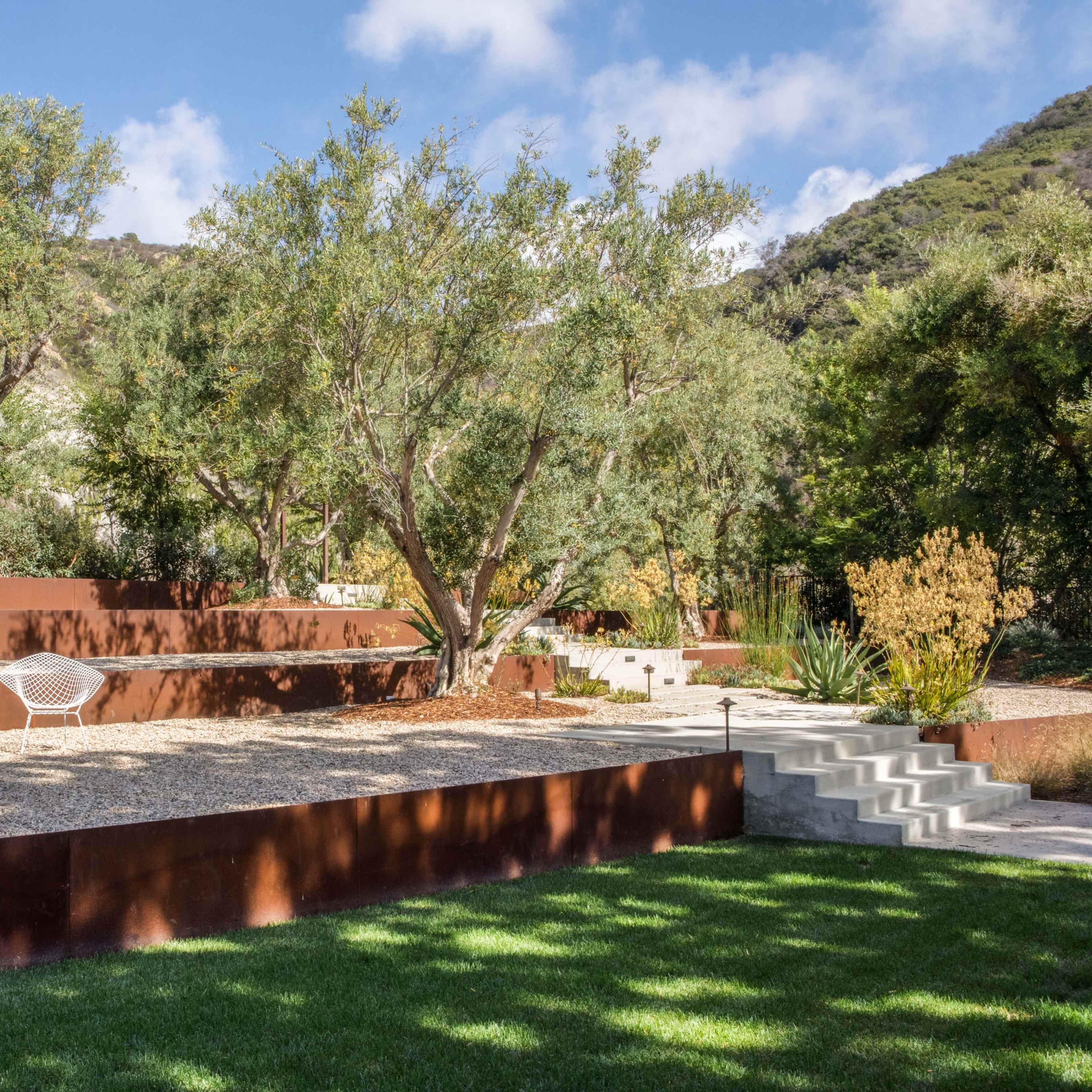 A landscaped garden featuring a gravel seating area with two white chairs, surrounded by rust-colored metal walls and various plants.