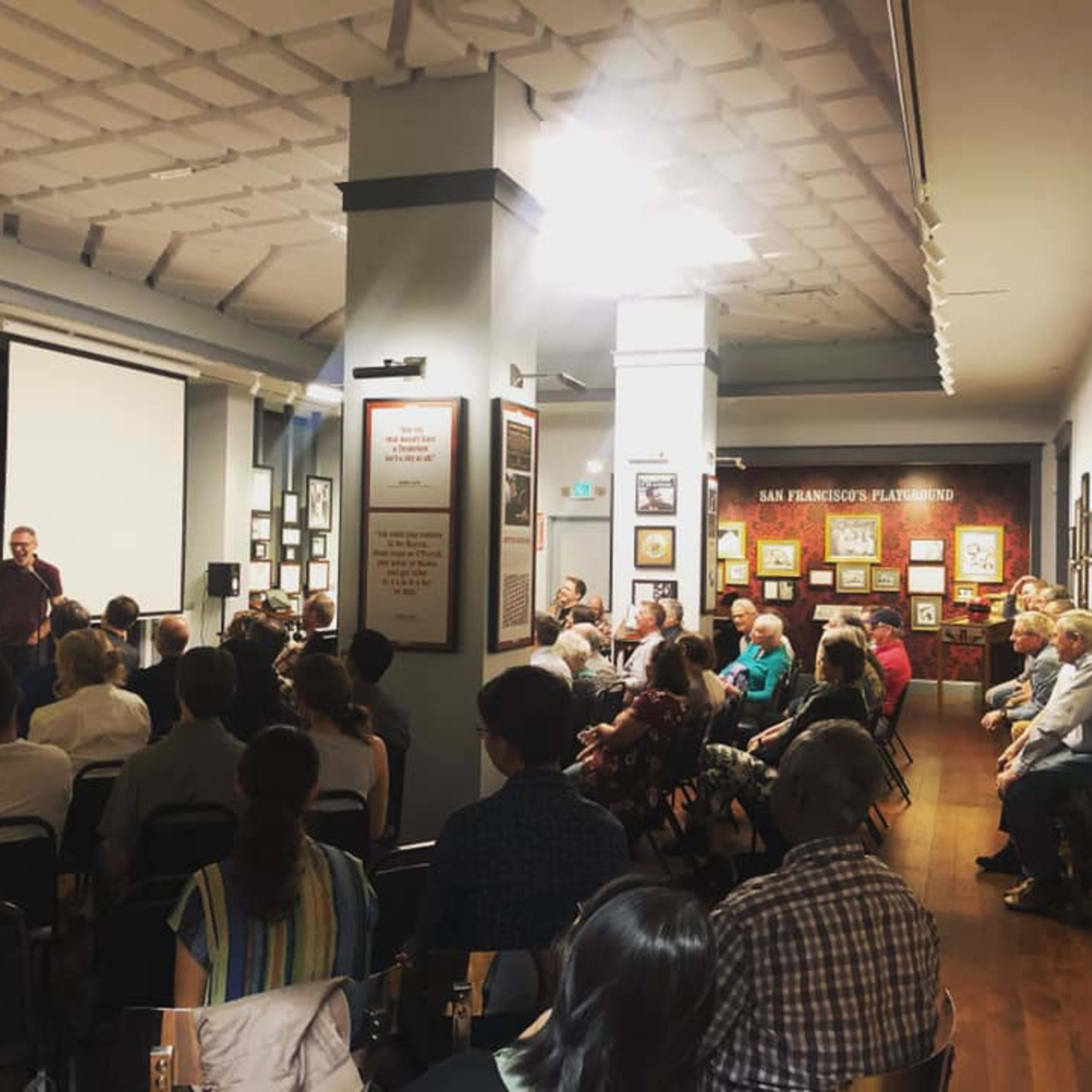 A crowd of people sits facing a speaker in a room adorned with framed photographs and artwork.