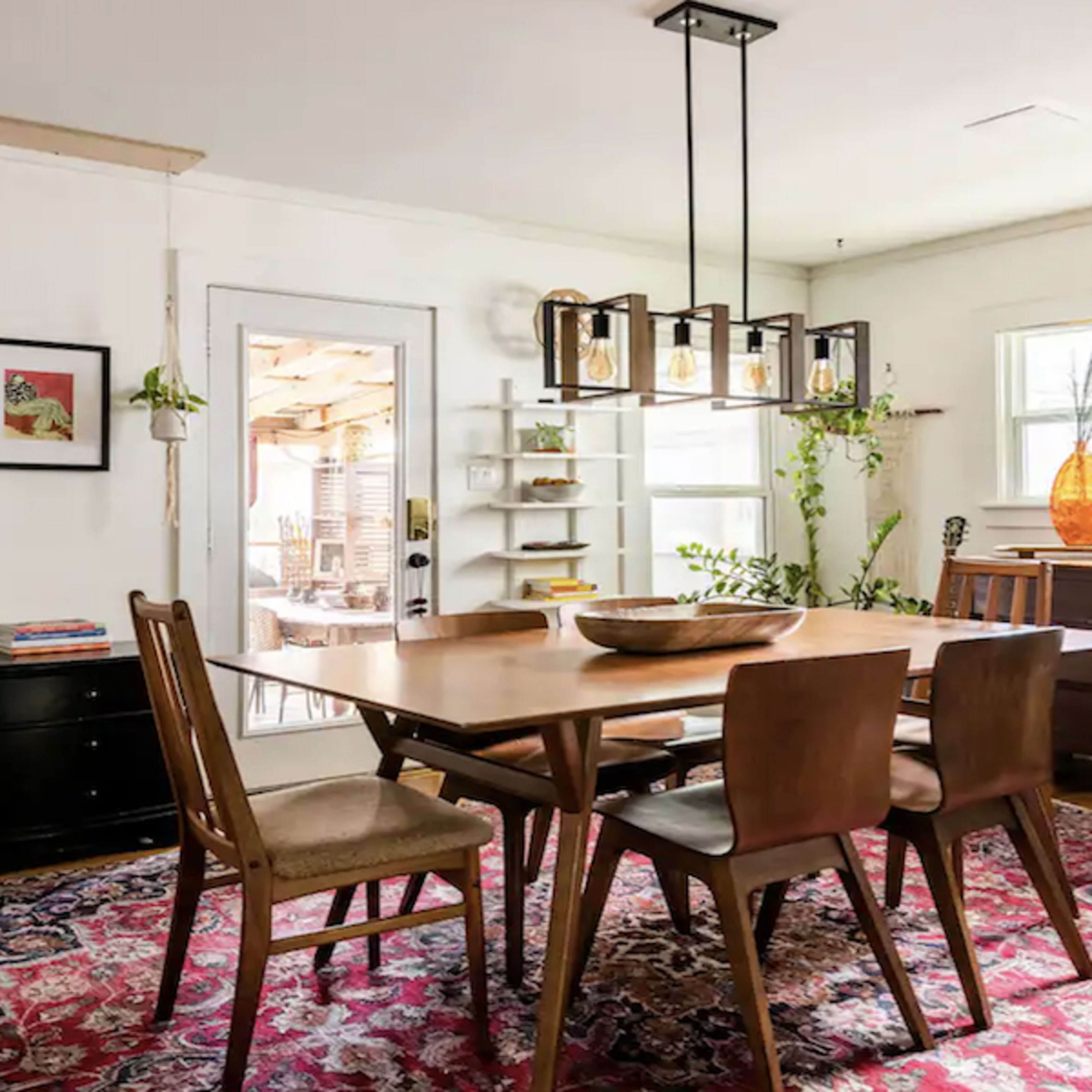 A dining area features a wooden table surrounded by chairs, with a light fixture overhead and large windows allowing natural light to enter.