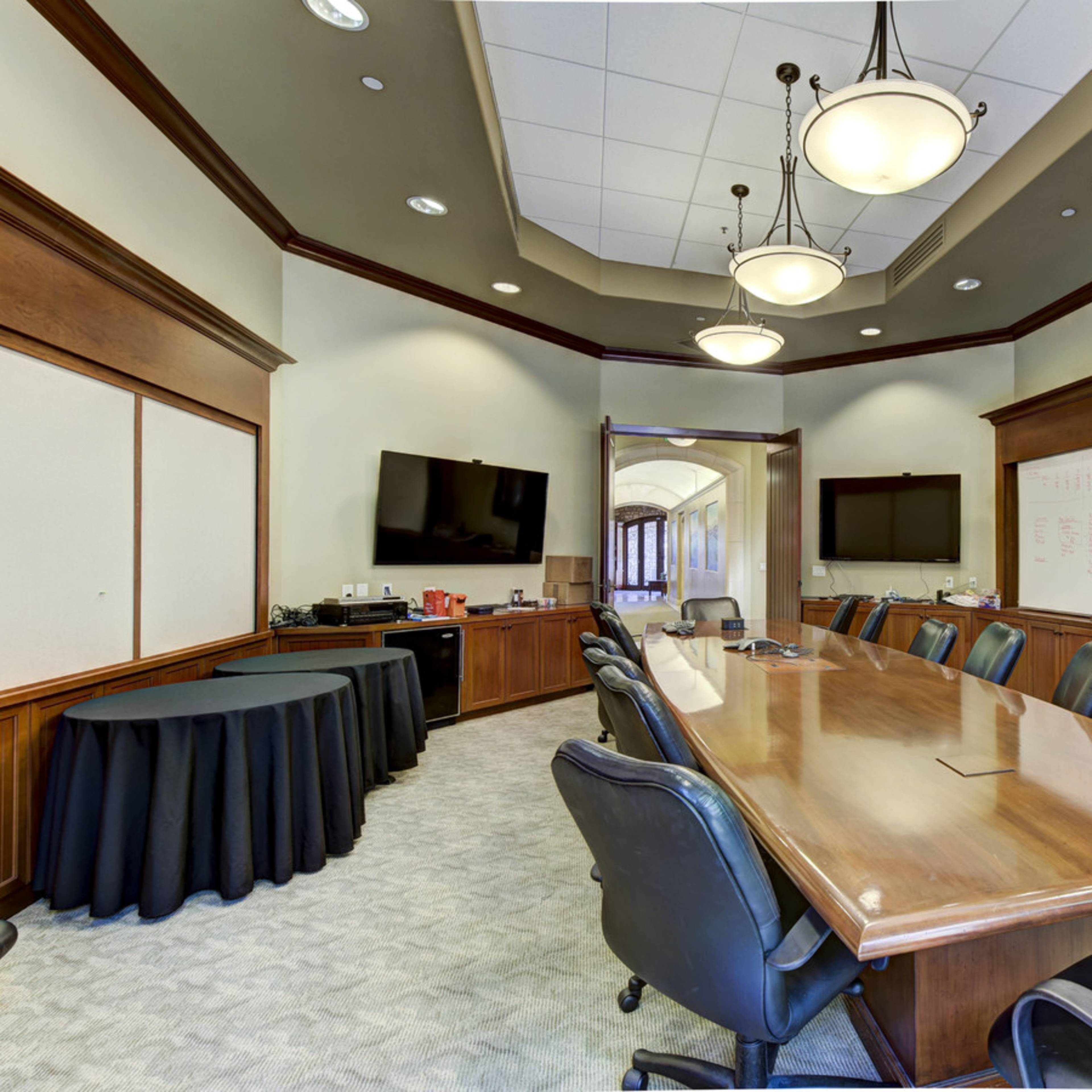 A large conference room with a long wooden table surrounded by black chairs and two flat-screen TVs mounted on the walls.