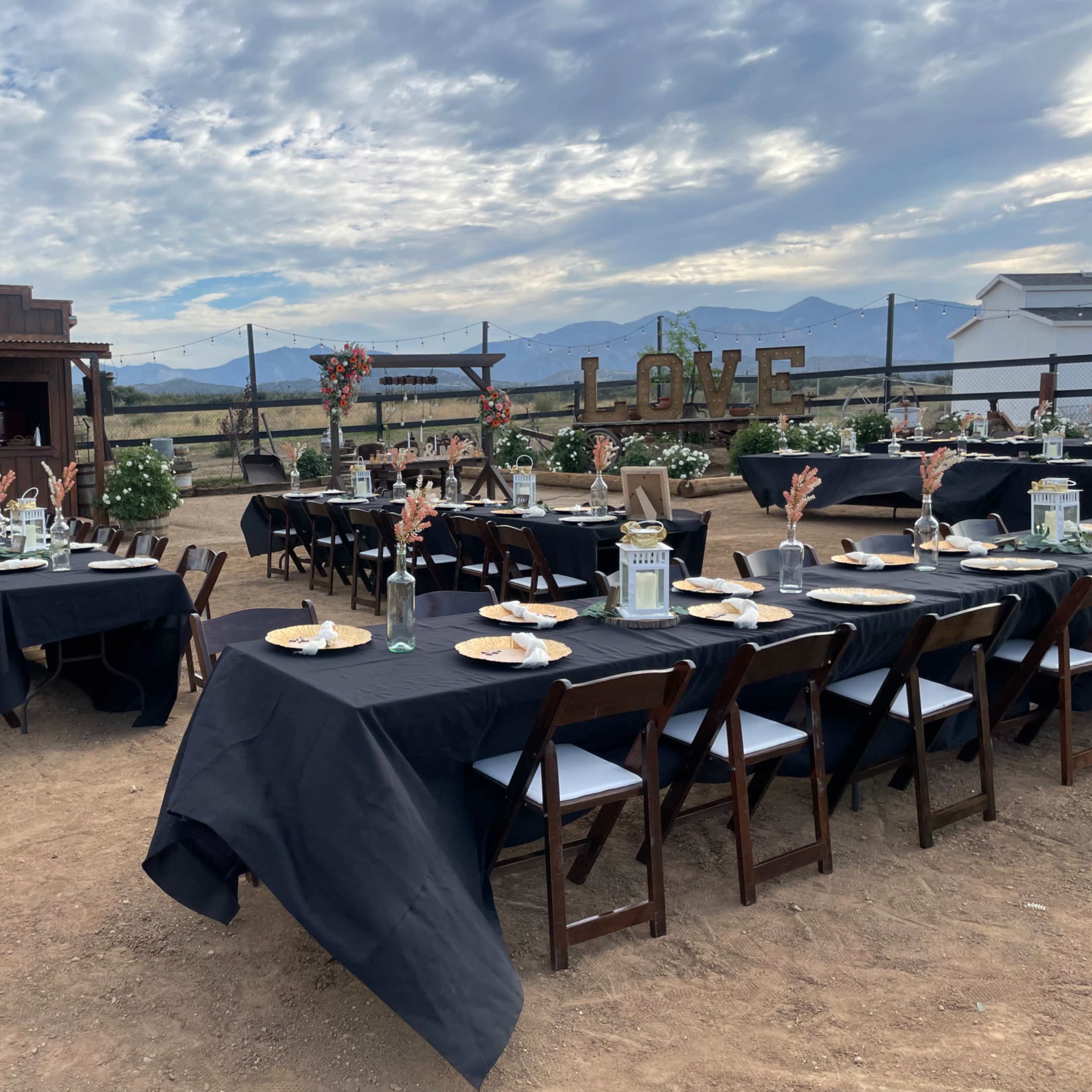 A long outdoor banquet table is set with black linens and decorative centerpieces, positioned in front of a rustic wooden structure under a cloudy sky.