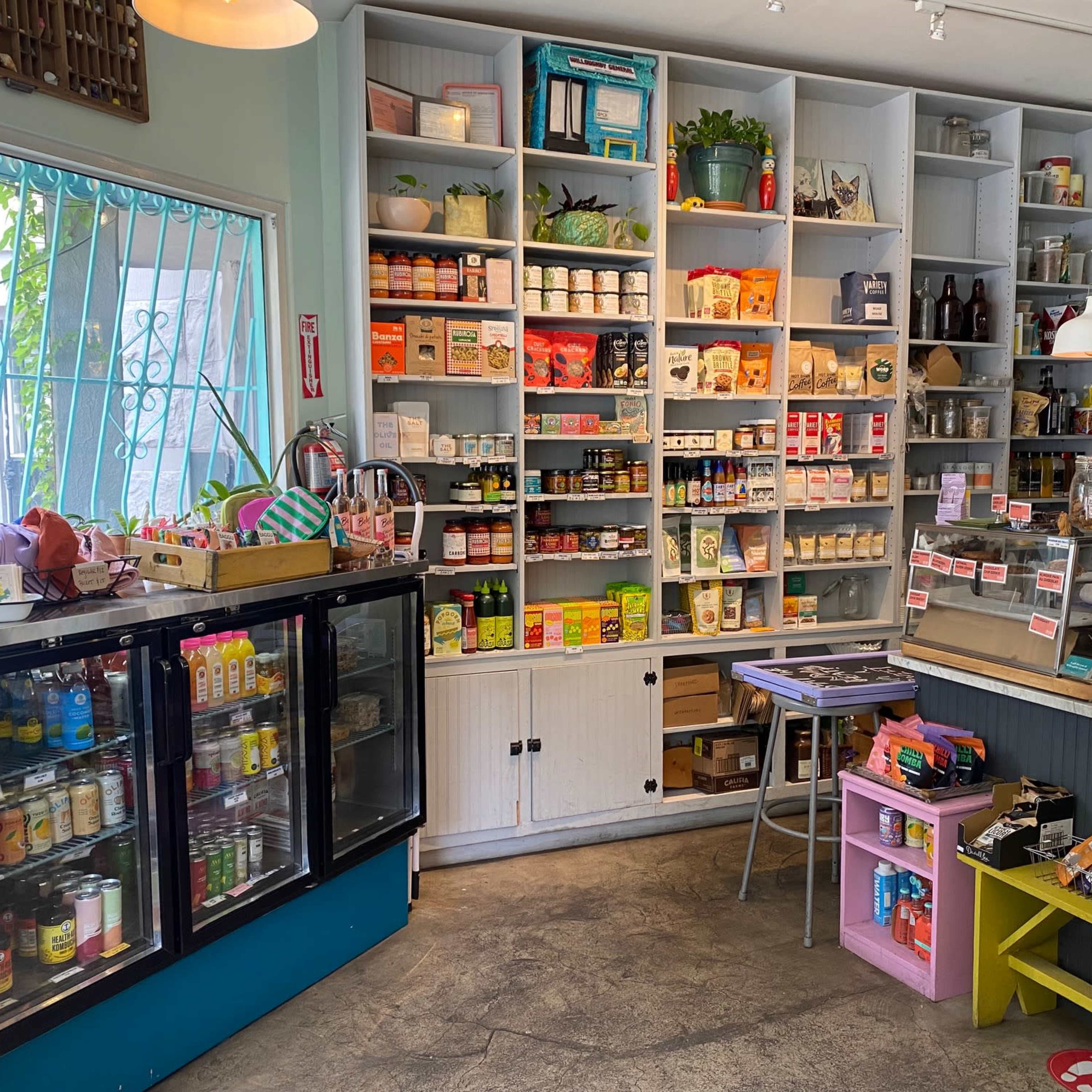 A small store interior featuring shelves stocked with various canned goods and products, a glass display refrigerator, and a counter with a cash register.