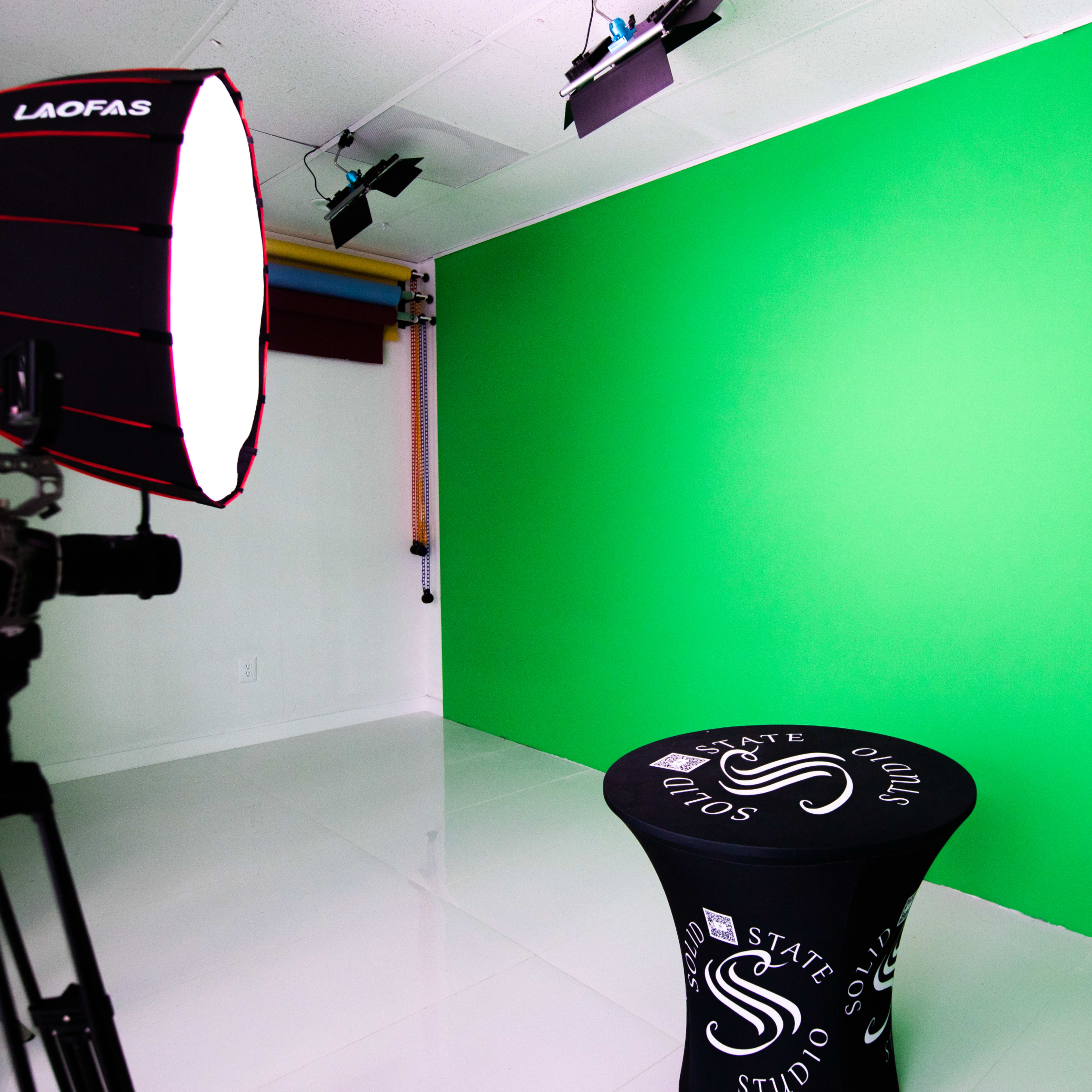 A studio setup featuring a green backdrop, a lighting umbrella, a camera on a tripod, and a table with a branded tablecloth.