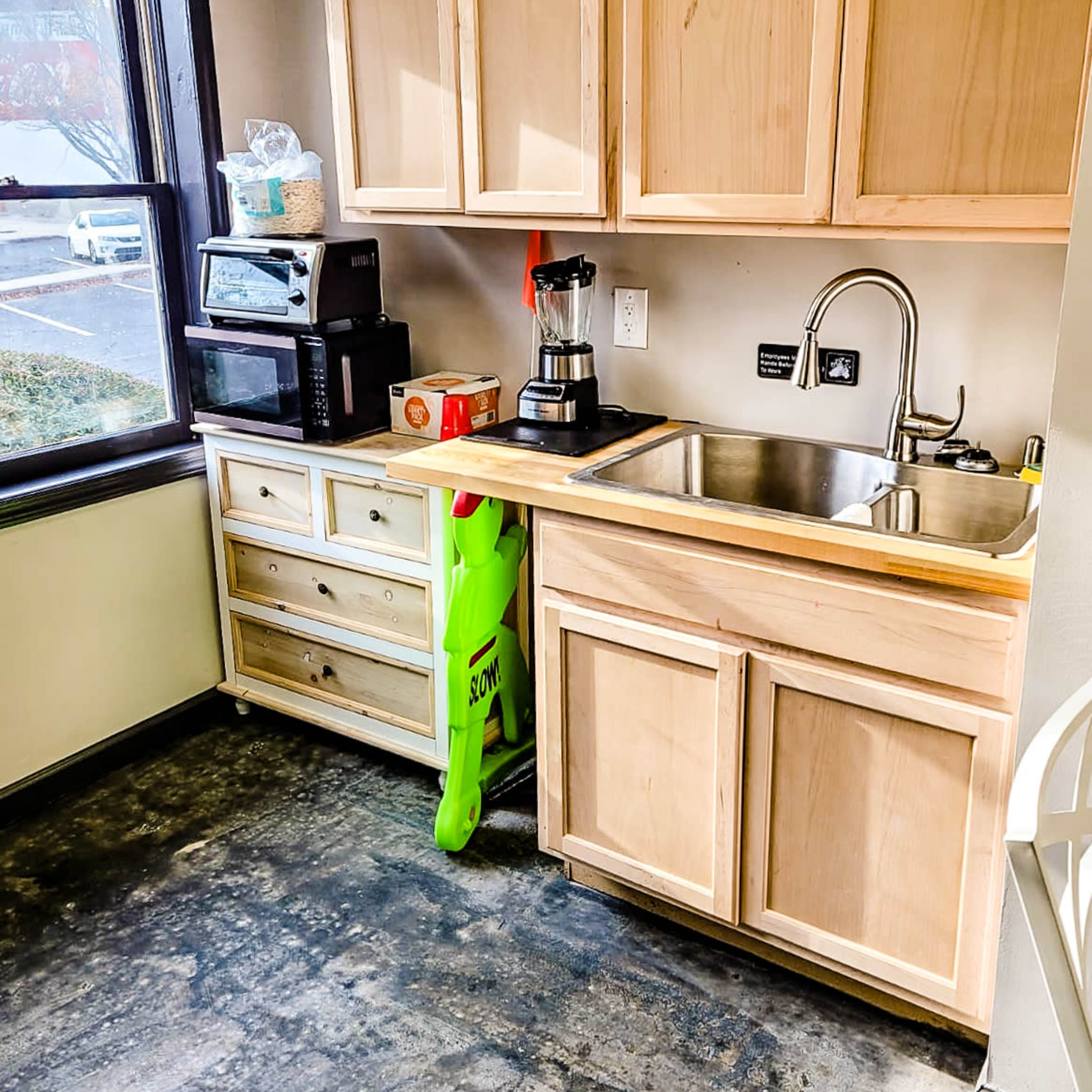 A kitchen area with wooden cabinetry, a stainless steel sink, a coffee maker, and a microwave on the counter, along with a green cleaning solution bottle on the floor.
