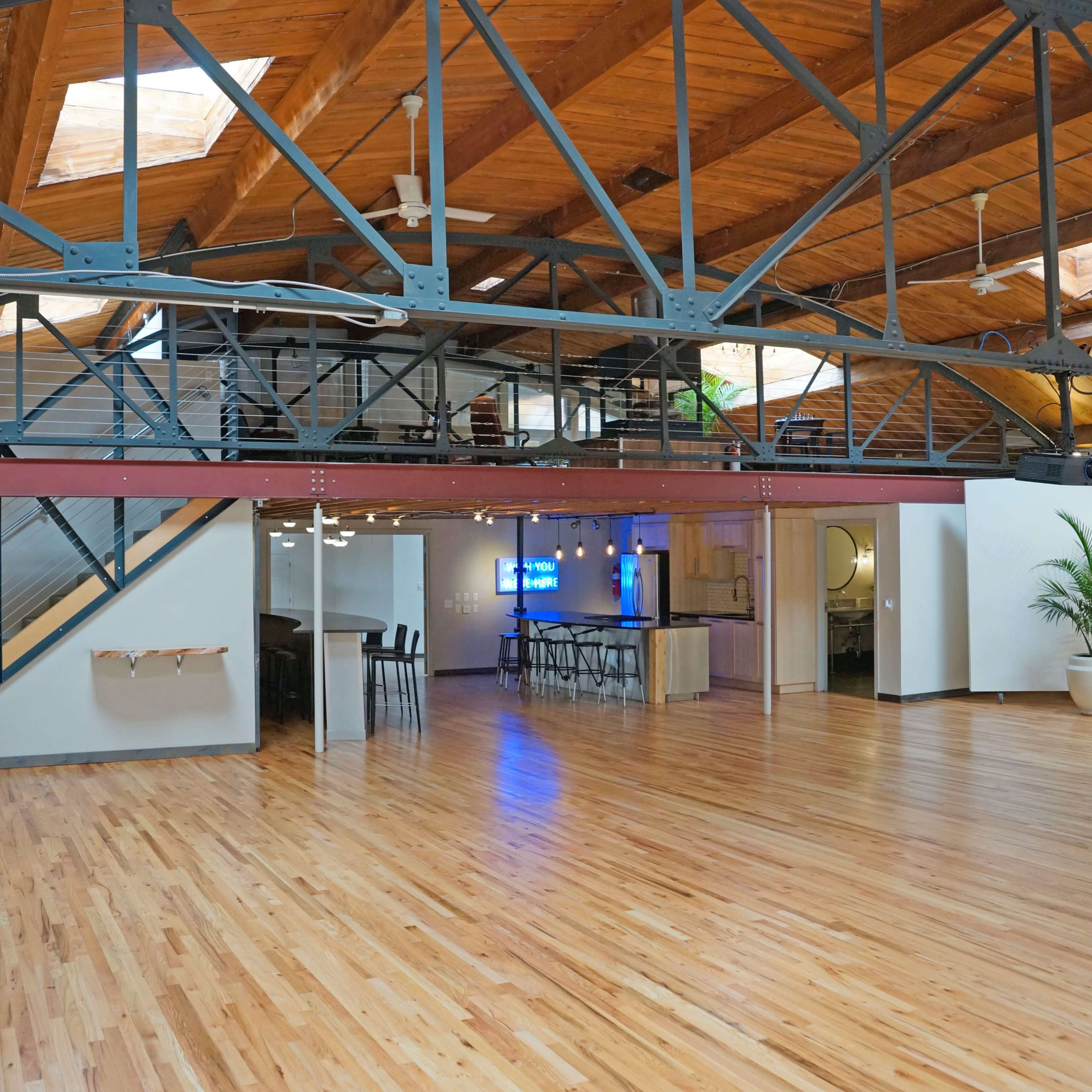An open-concept space with a wooden floor, exposed beams, and a staircase leading to a second level, featuring a bar area in the background.