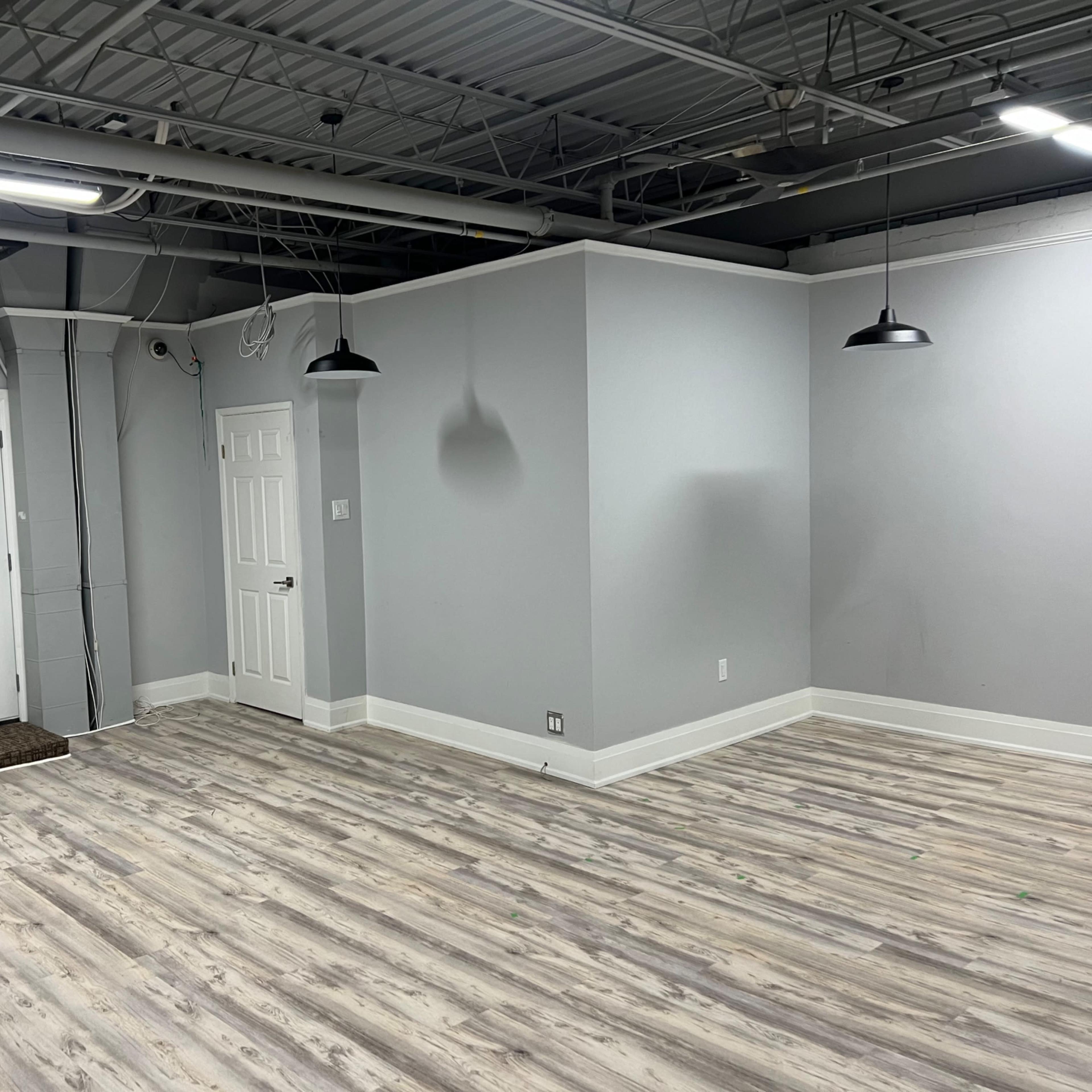 A partially empty, light gray room with two doors, hanging lights, and wooden flooring.