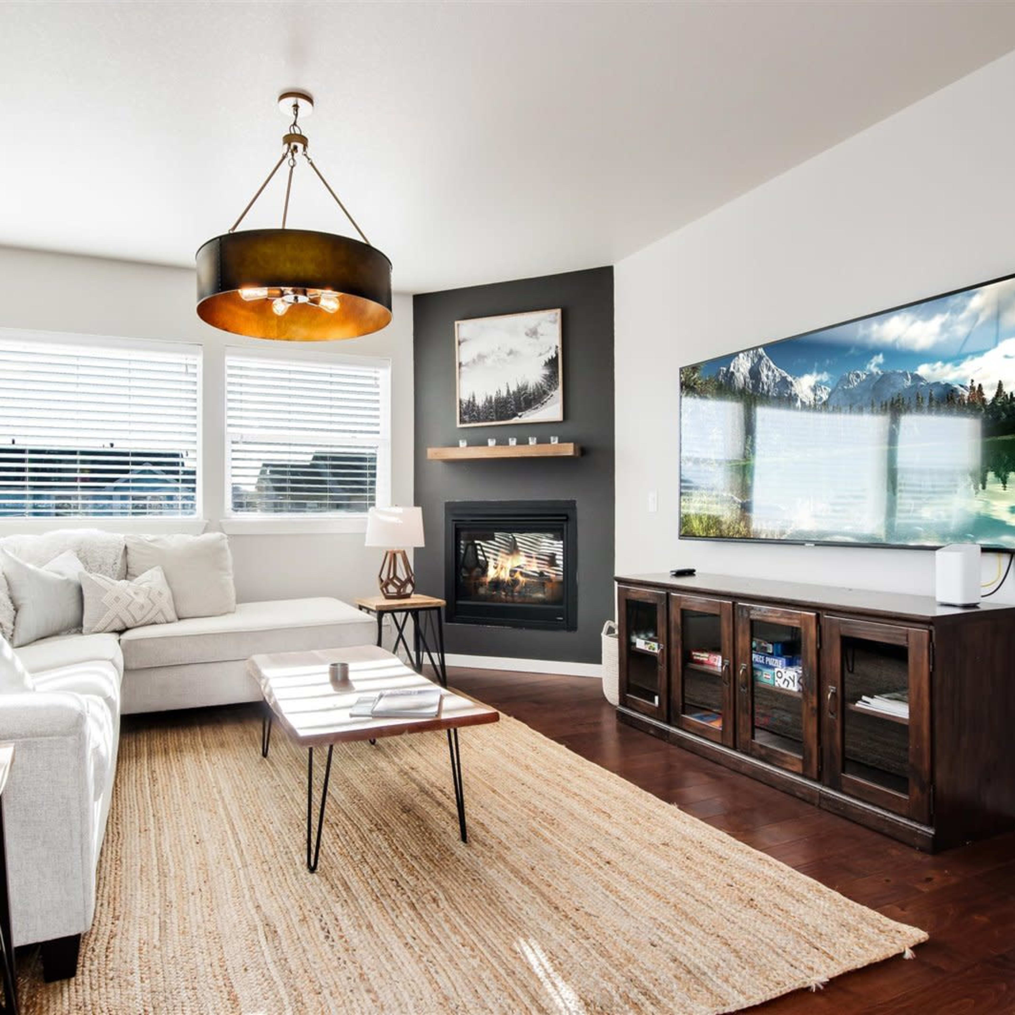 A modern living room features a sectional sofa, a coffee table, a TV mounted on the wall, and a fireplace, with large windows allowing natural light in.