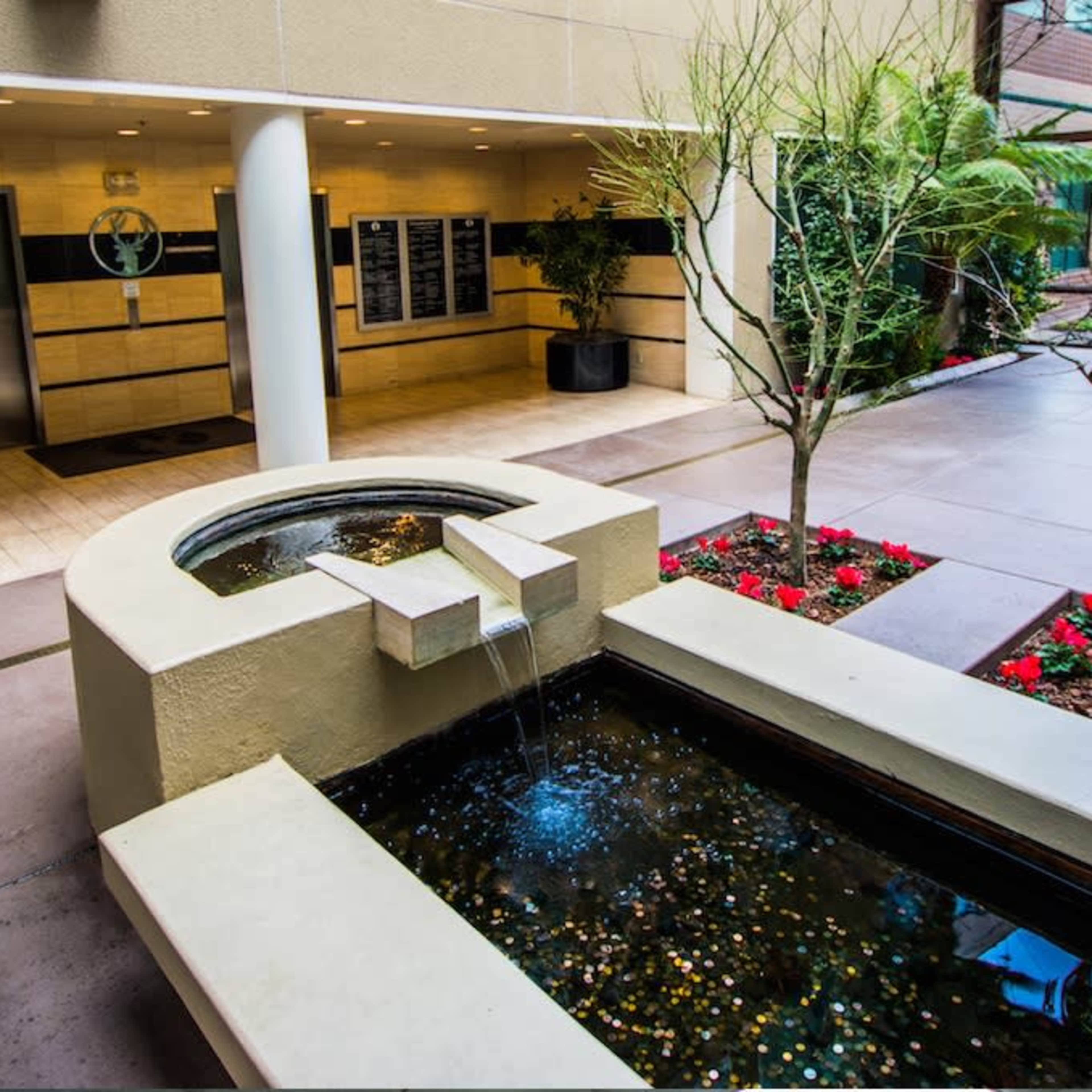 A modern indoor space featuring a water fountain surrounded by plants and red flowers, with an elevator in the background.