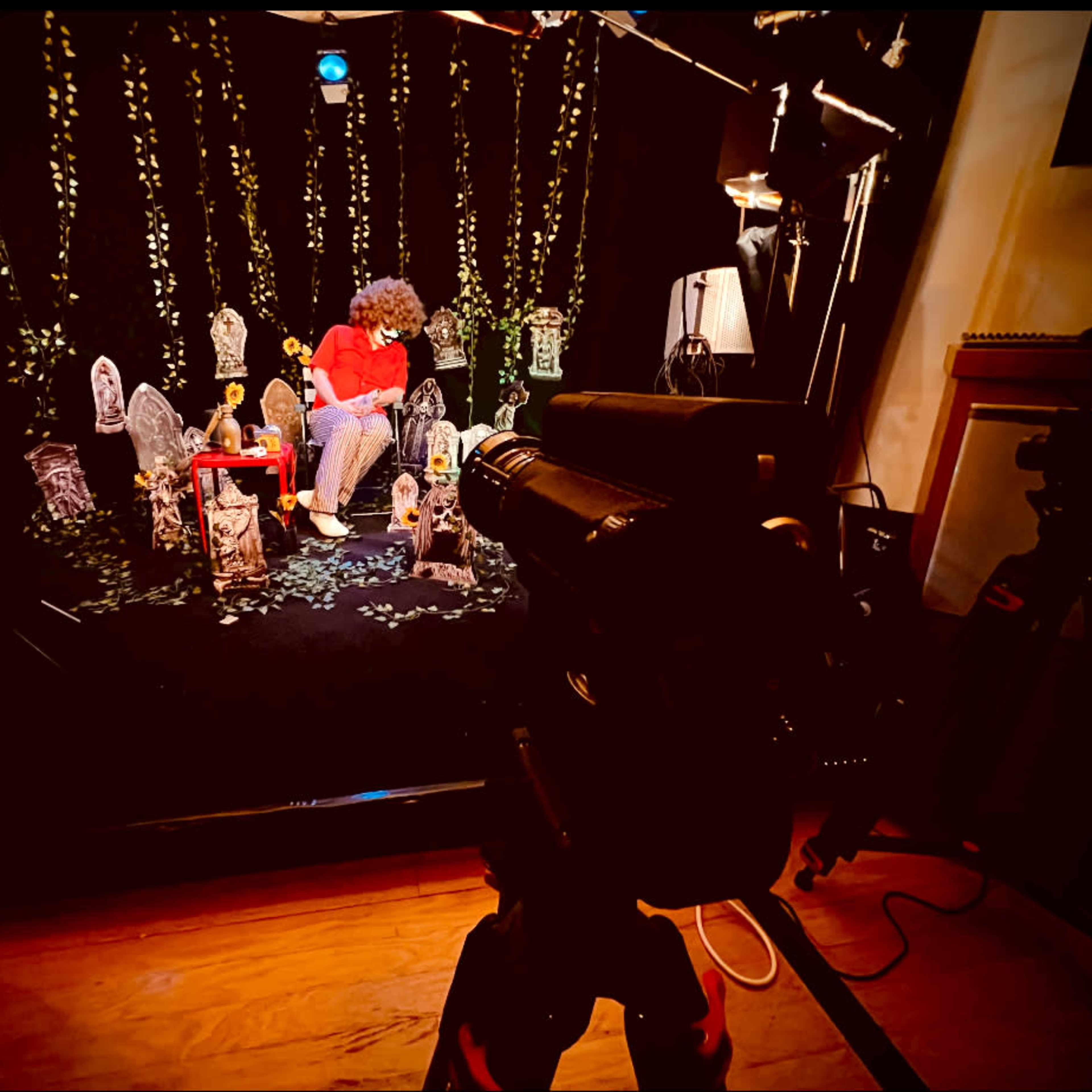 A person sits on a set decorated with greenery and various props, while a camera is positioned in the foreground.
