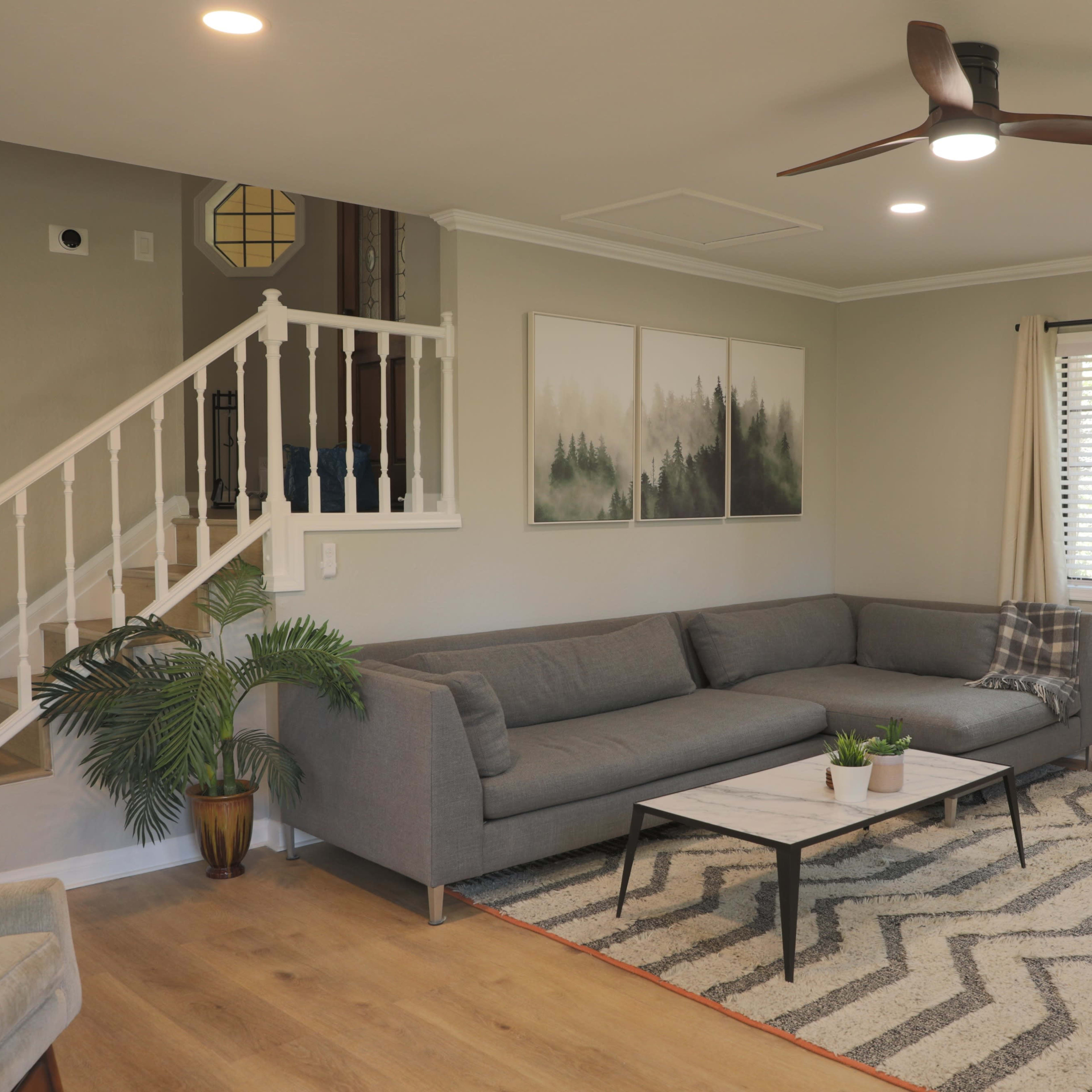 A modern living room features a gray sectional sofa, a coffee table on a patterned rug, and large windows with curtains.